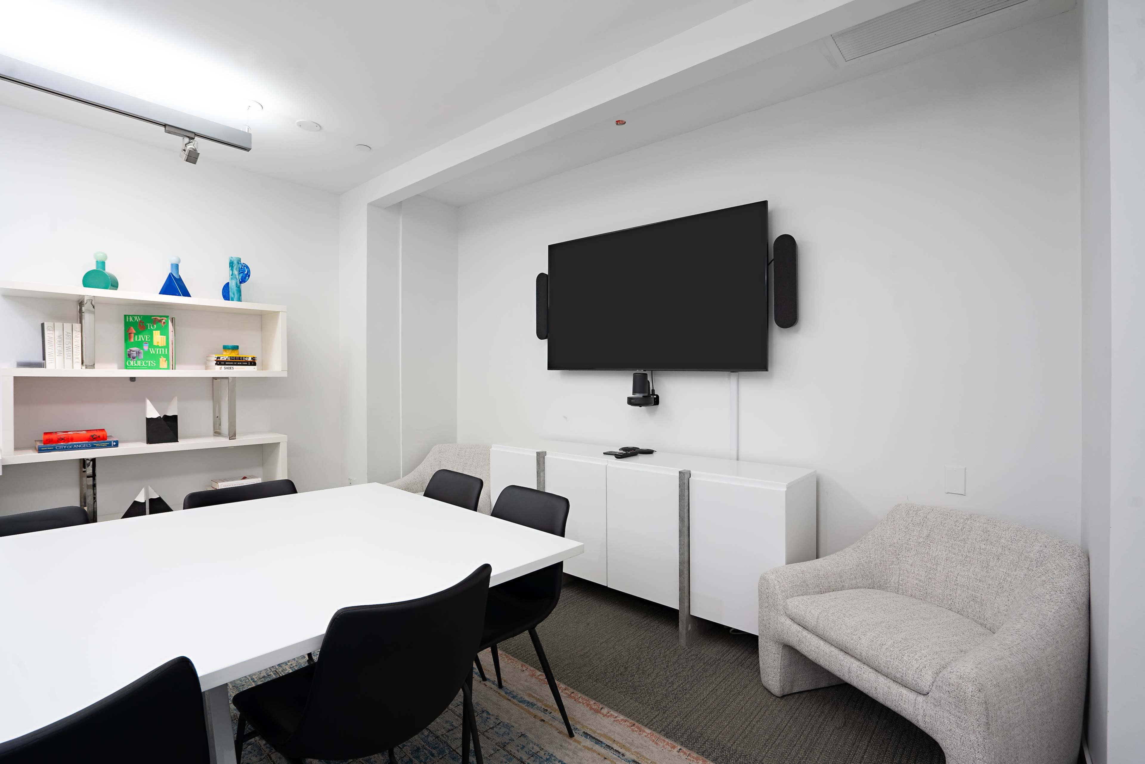 A modern conference room features a white table surrounded by black chairs, with a television mounted on the wall and a white storage unit displaying decorative items.