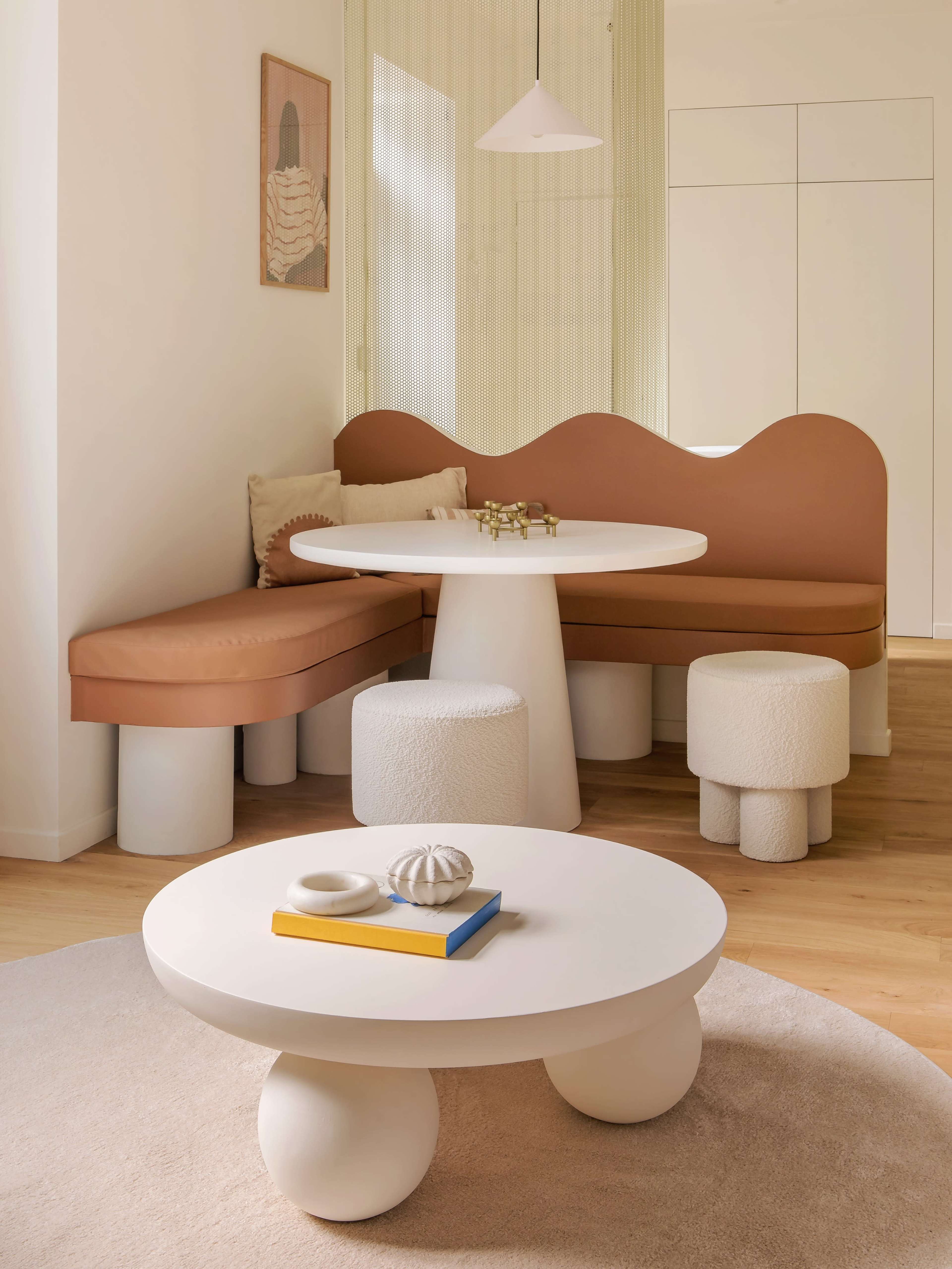 A modern interior features a round table and seating area with a wavy backrest, complemented by cylindrical stools and minimal decor.
