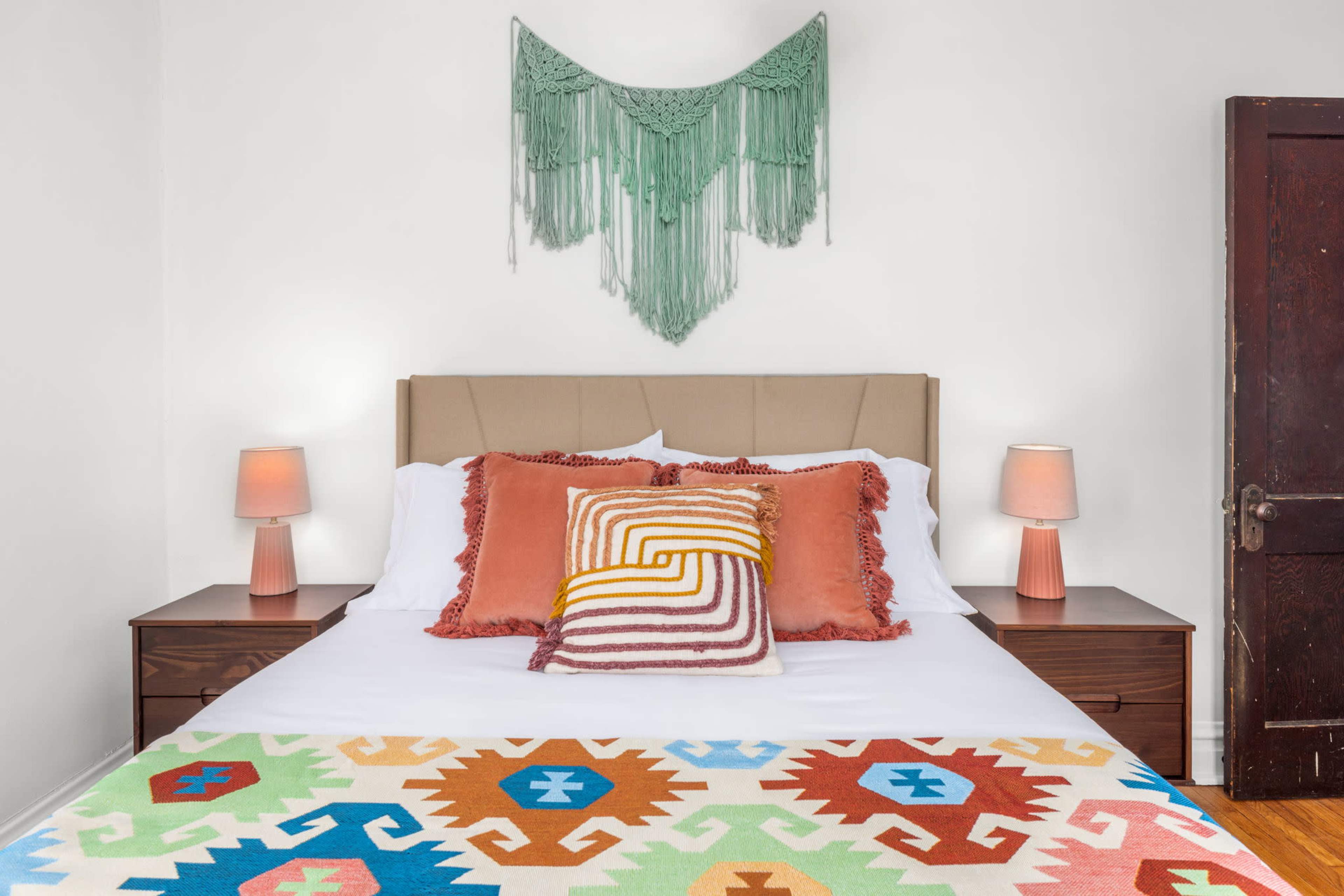 The image shows a bedroom featuring a bed with white linens, colorful pillows, and a patterned throw, flanked by two bedside tables with lamps, and adorned with a macramé wall hanging above the headboard.
