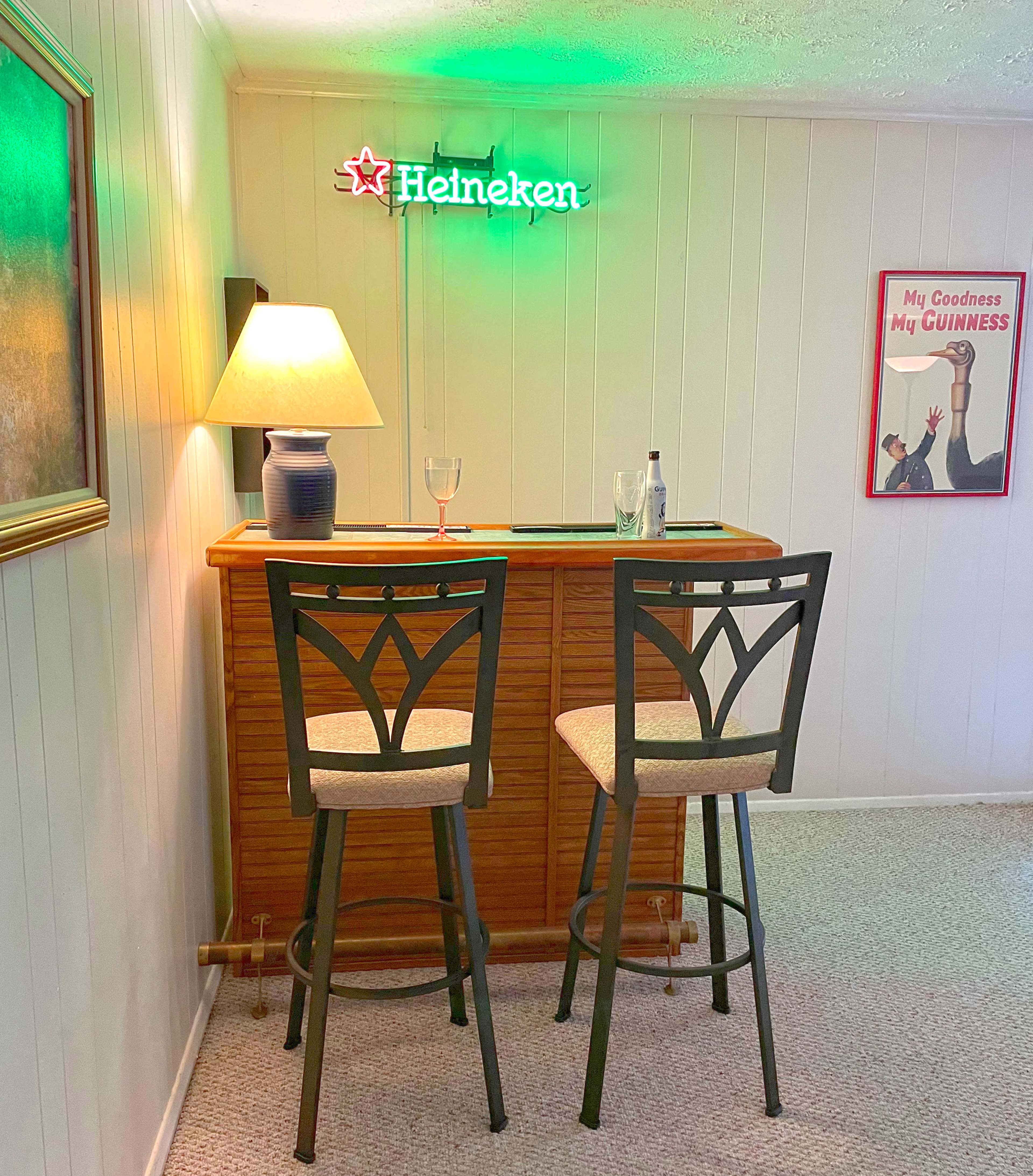 A small home bar area with two high stools, a lamp, a Heineken neon sign, and a framed Guinness poster on the wall.