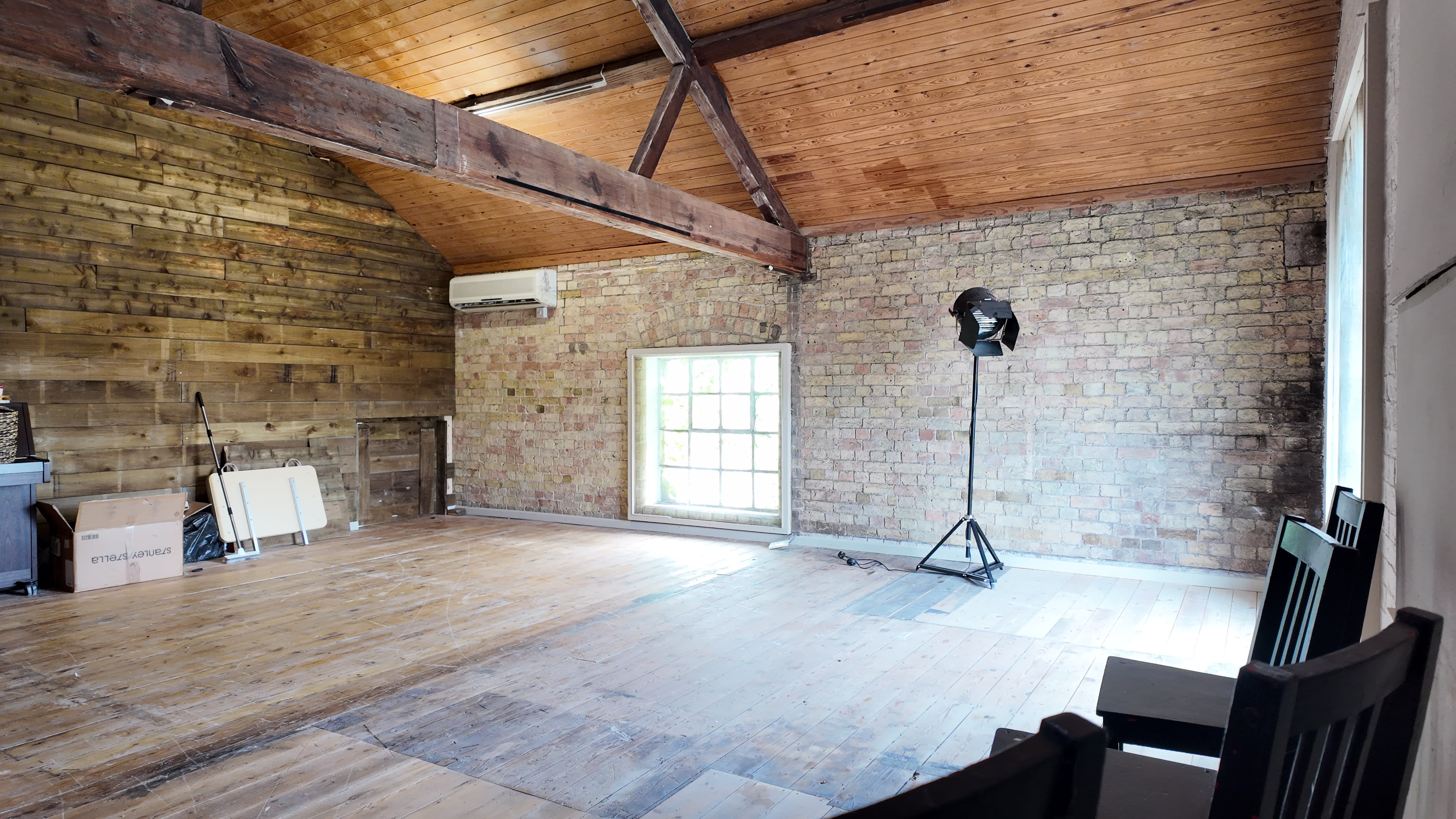 The image shows a spacious, unadorned room with wooden floors, a tall window, exposed brick walls, and a light stand positioned near the window.