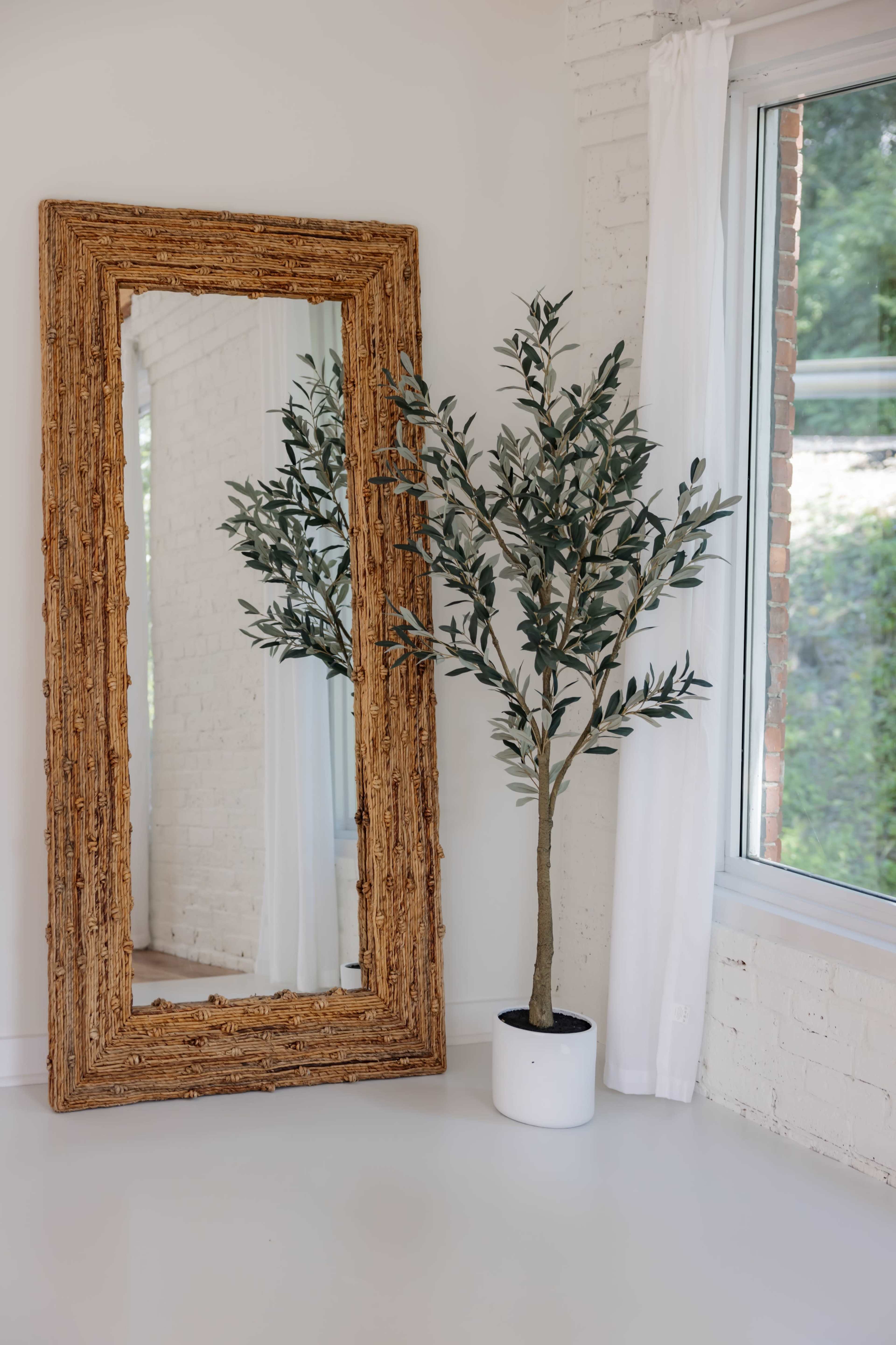 A large, textured mirror stands against a white wall next to a potted faux olive tree, with sunlight streaming in through a nearby window.