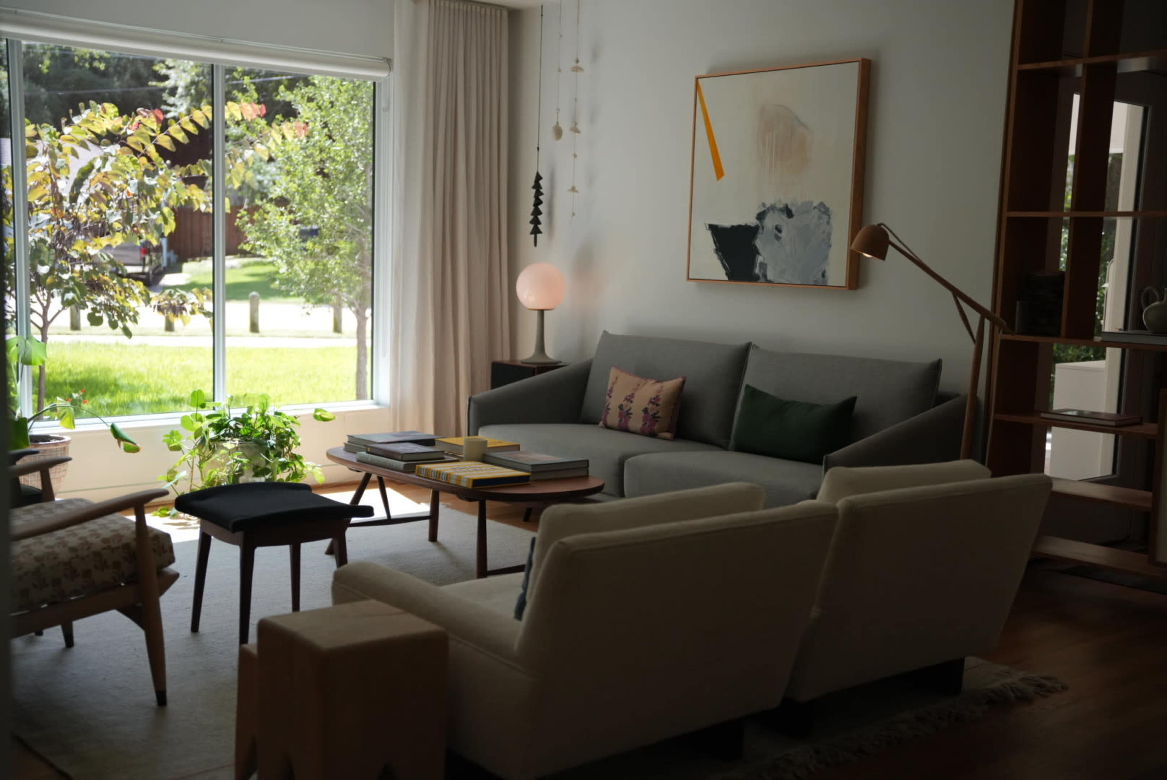 A cozy living room features a gray sofa, a wooden coffee table stacked with books, and a large window displaying greenery outside.