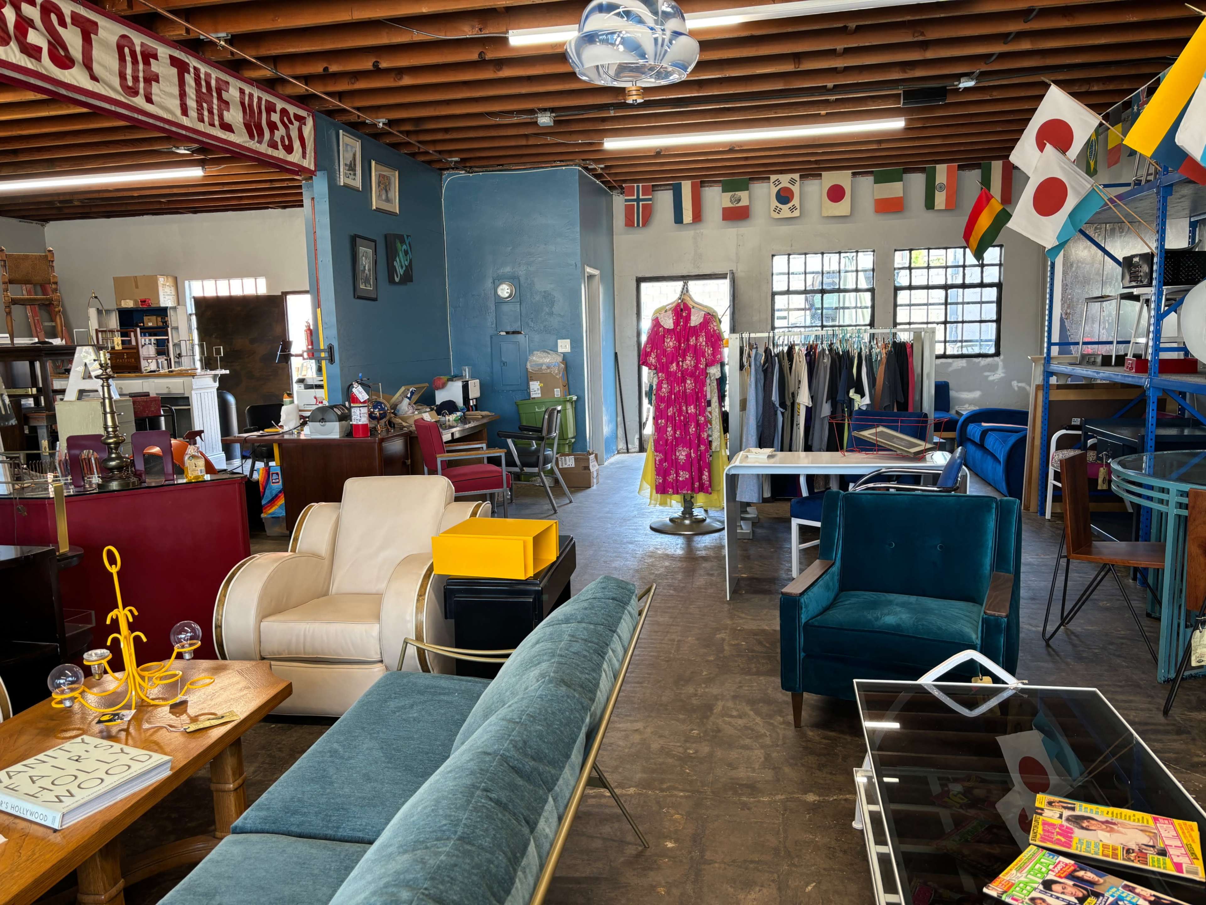 A brightly lit thrift store with various pieces of furniture, including chairs, a sofa, and clothing racks adorned with different national flags.