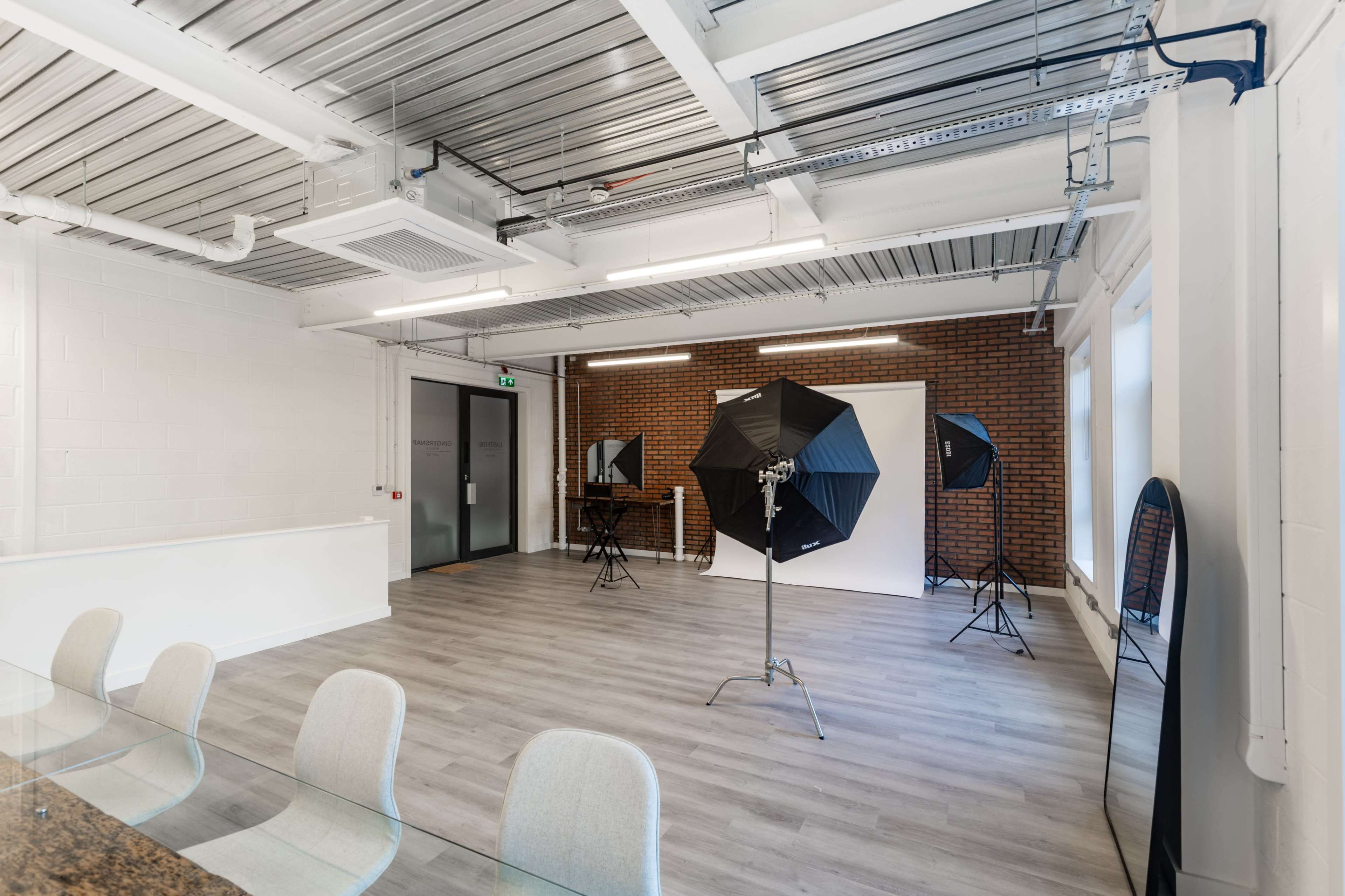 The image shows a spacious photography studio with a white backdrop, lighting equipment, and a large table in a well-lit area.