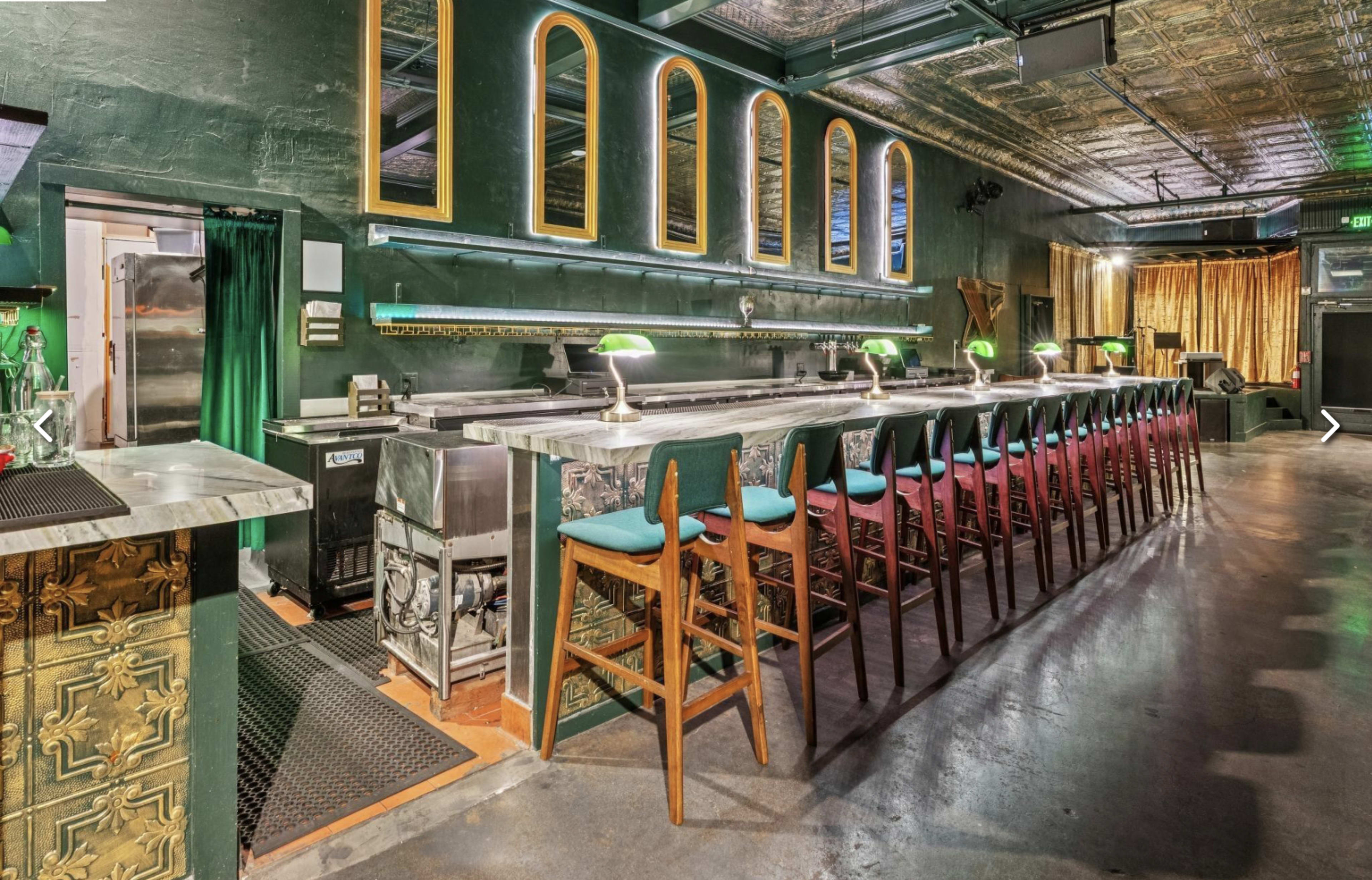 The image shows a bar area with a long counter, high stools, green walls, and decorative mirrors, illuminated by soft lights.