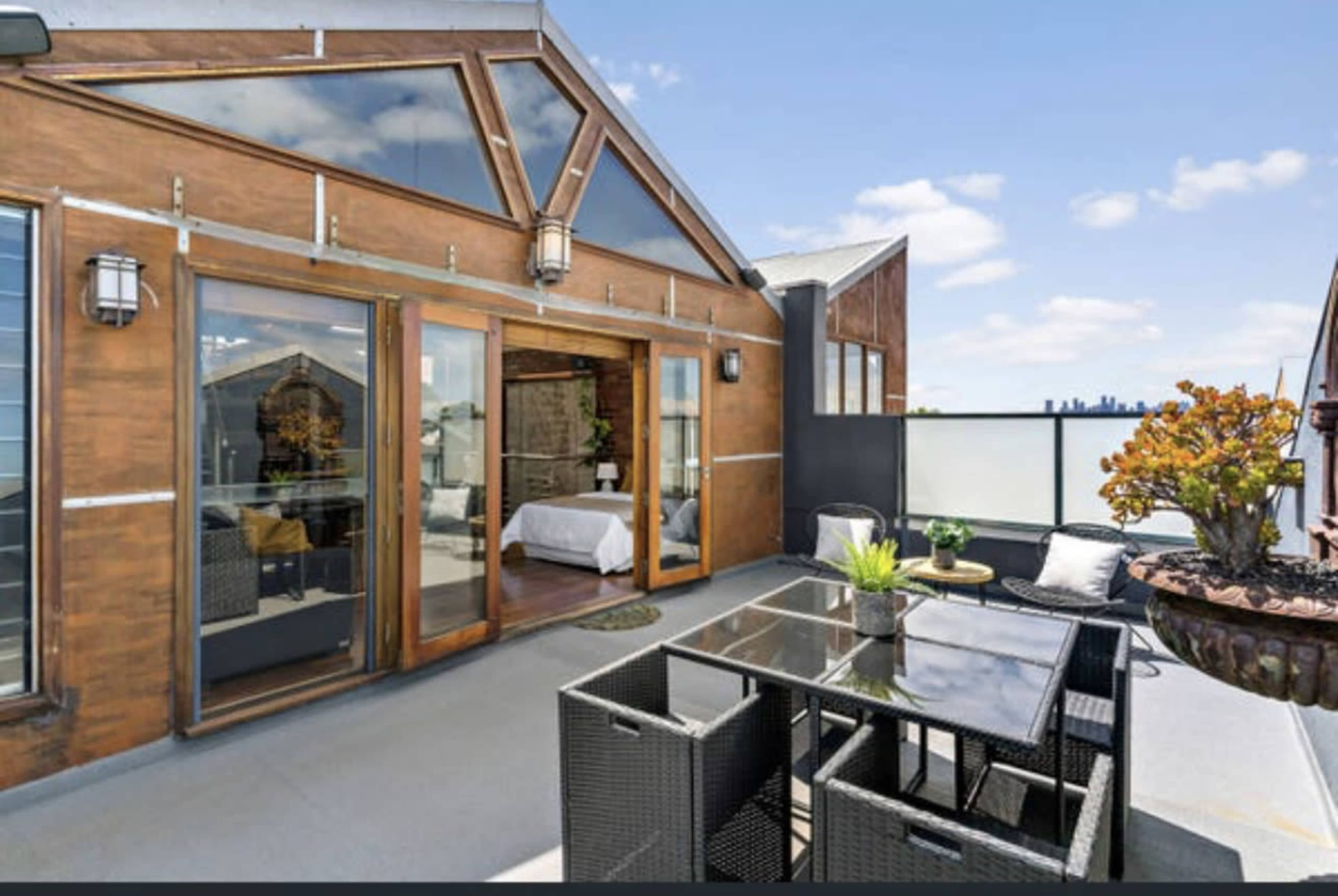 A modern rooftop patio featuring a glass table surrounded by black wicker chairs, with sliding glass doors leading into an interior space.