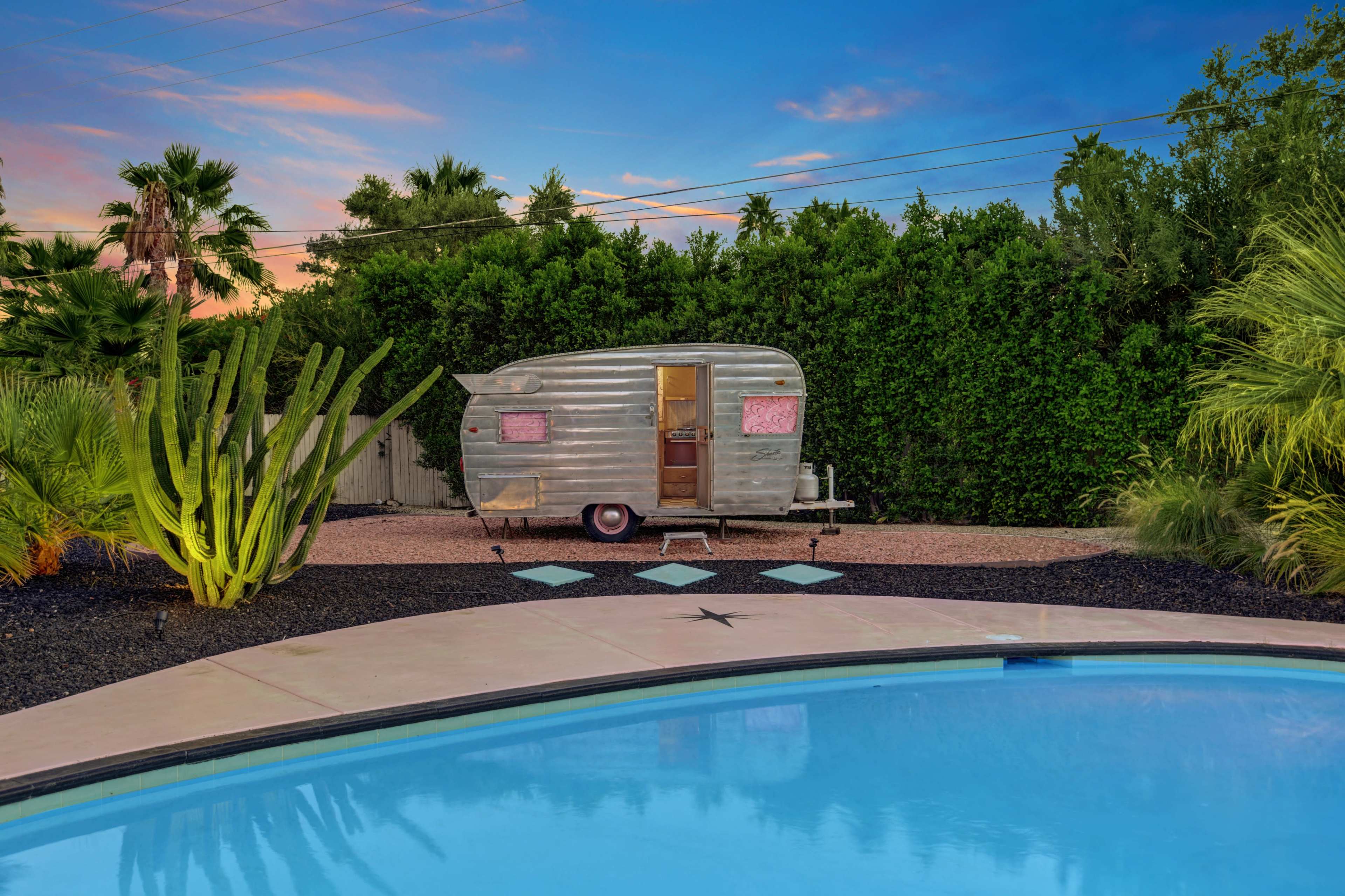 A vintage silver camper is parked beside a swimming pool surrounded by tropical plants under a colorful sky at dusk.