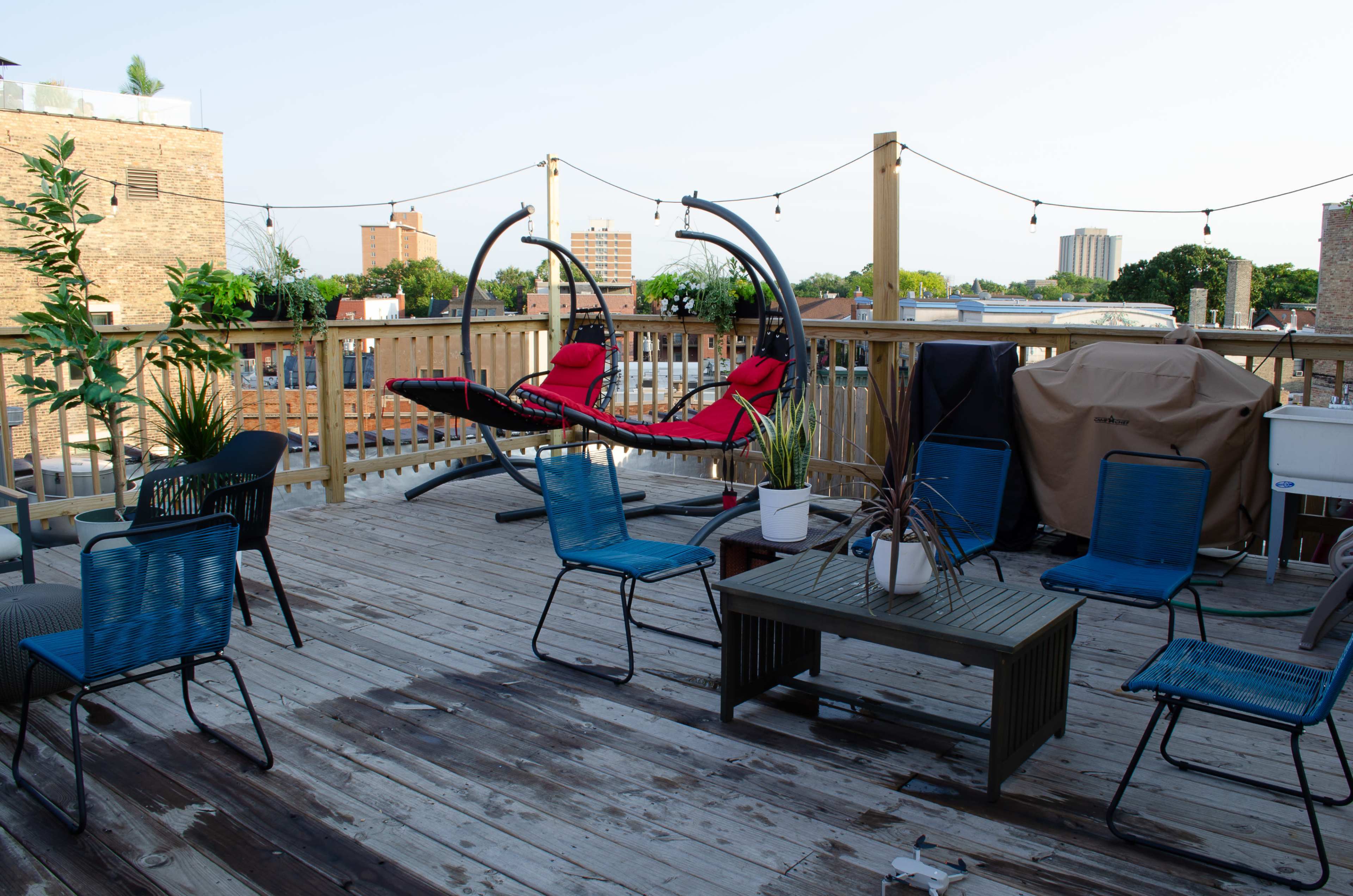 A wooden rooftop deck furnished with several chairs, a coffee table, and a hanging chair, surrounded by potted plants and city buildings in the background.