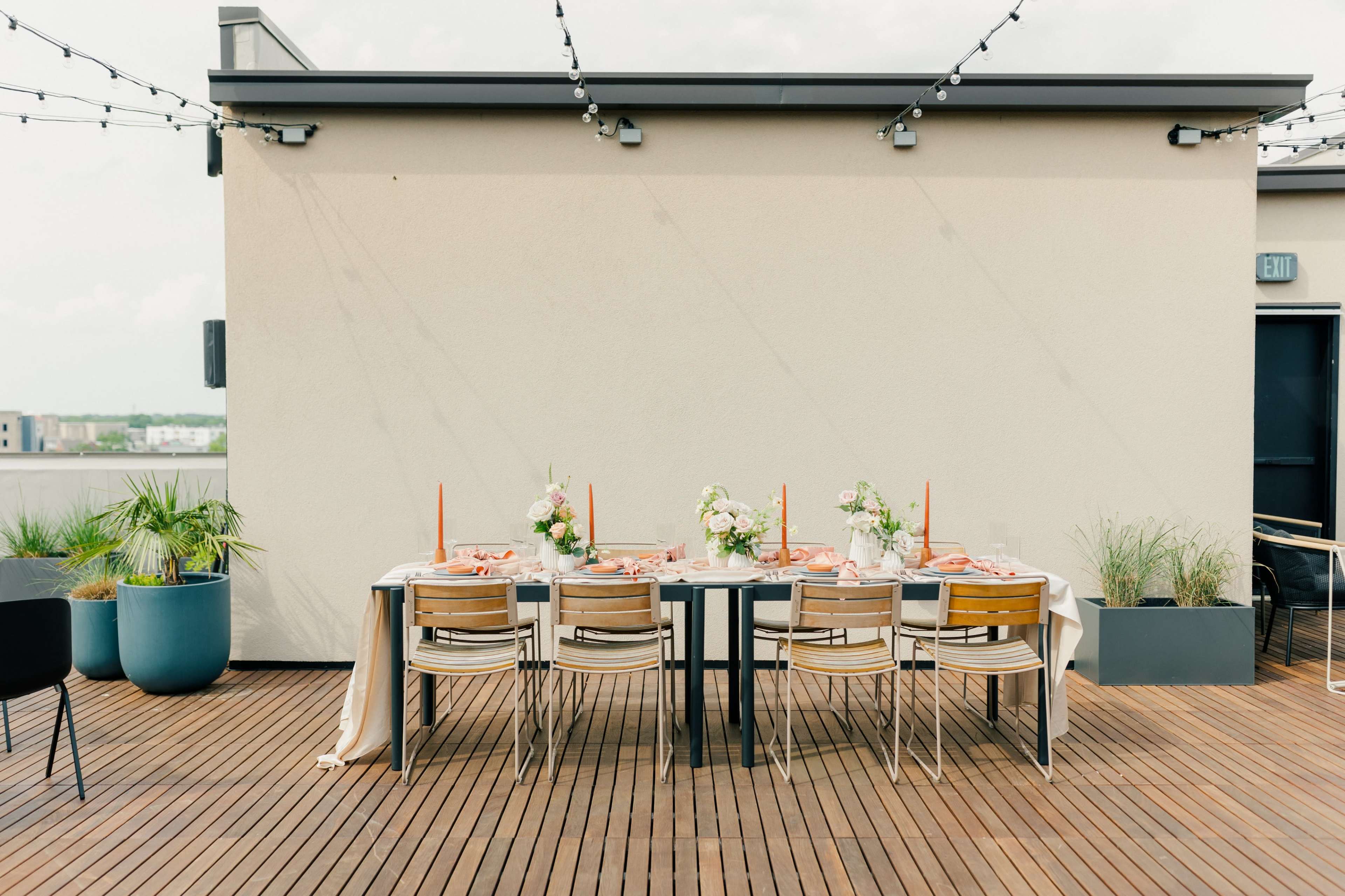 A long dining table set for a meal with floral centerpieces and candles is positioned on a wooden deck overlooking a cityscape.