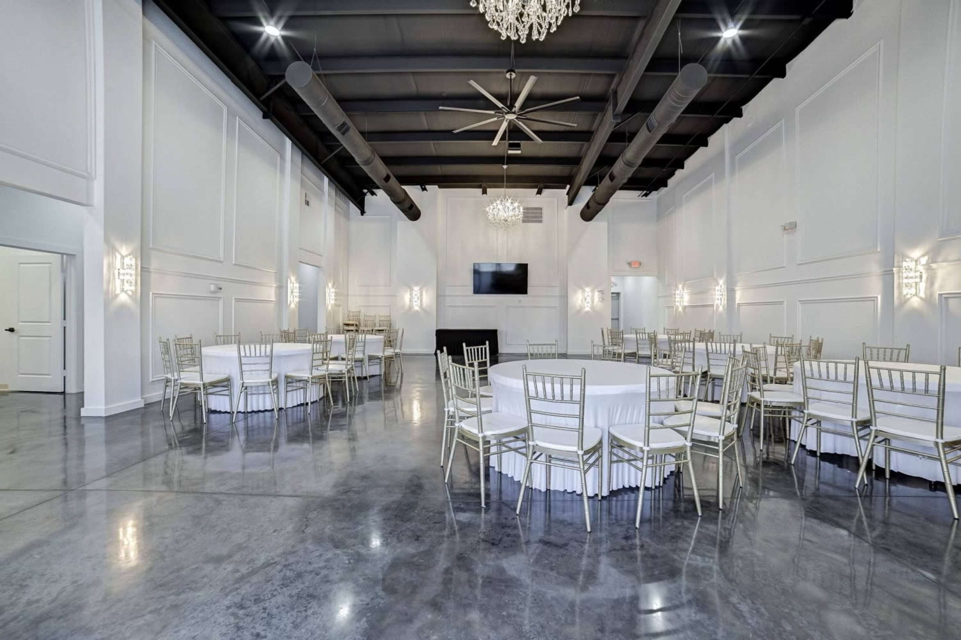 The image shows an elegantly designed event space with round tables, white tablecloths, and a polished concrete floor, illuminated by modern chandeliers.