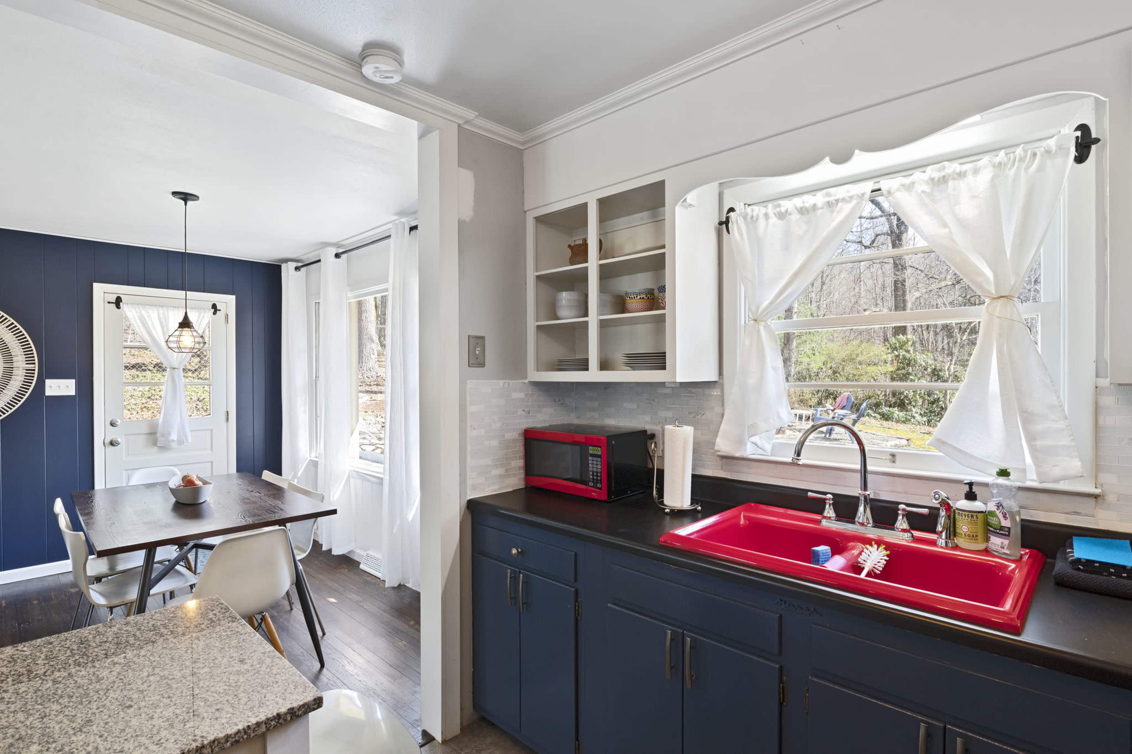 A kitchen with a red sink and countertop, white cabinets, and a dining area visible through the window.