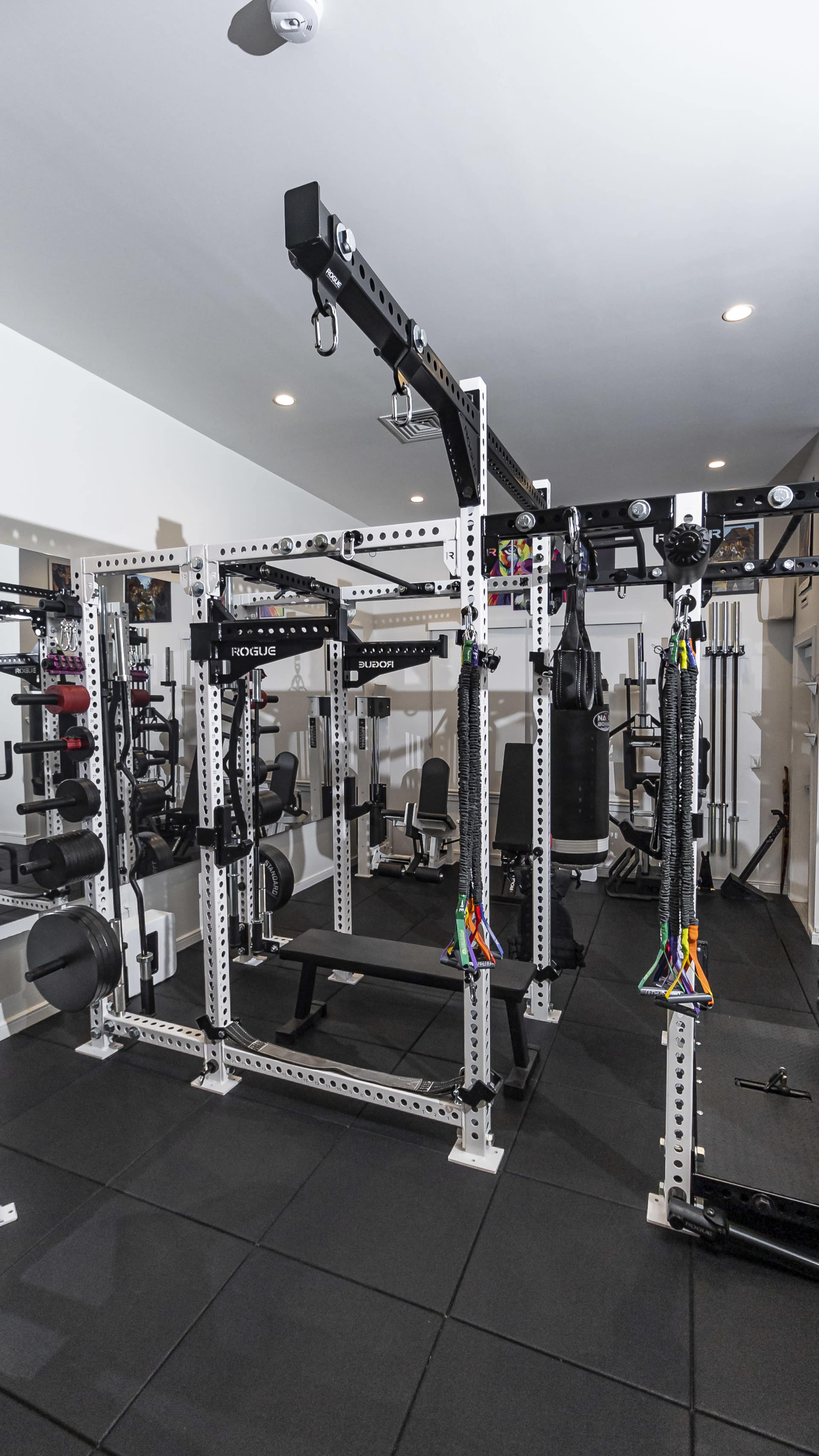 A well-equipped home gym featuring a multi-station weight rack, exercise machines, and various weights on a rubber flooring surface.