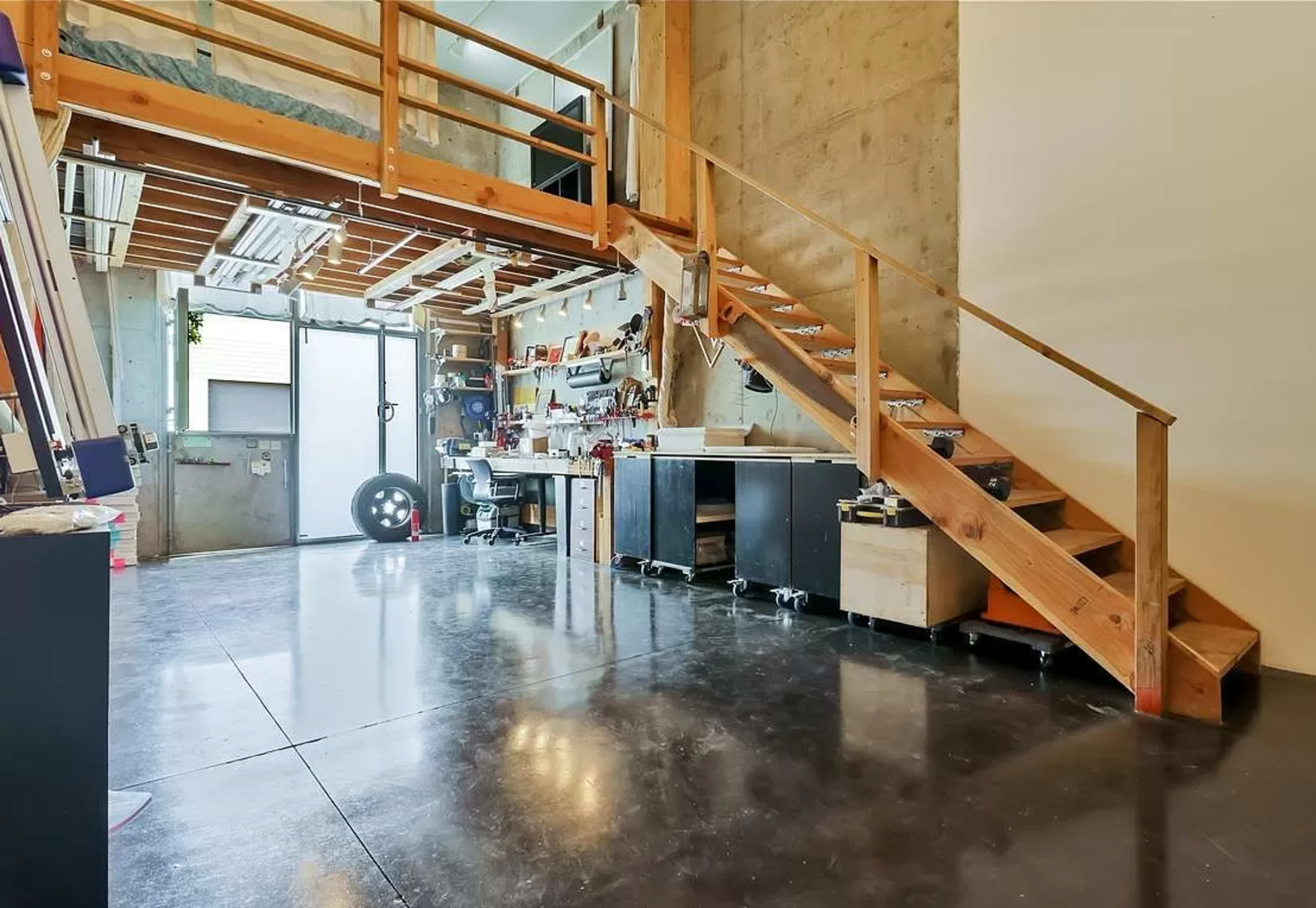A two-level workspace with a wooden staircase leading to a lofted area, featuring a glass door and various tools and equipment on the ground level.