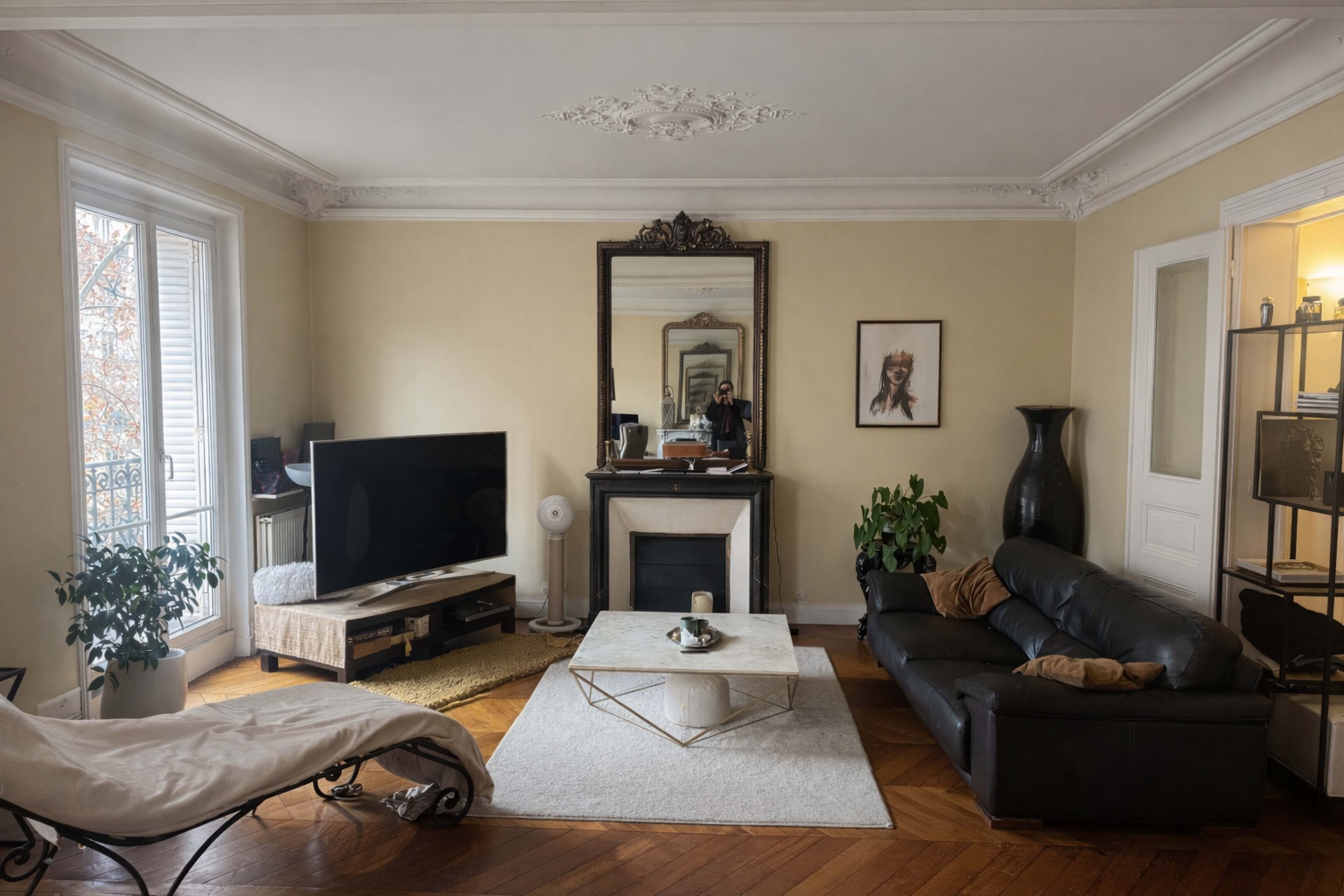 The image shows a living room featuring a black leather sofa, a coffee table, a large mirror above a fireplace, and a television on a wooden stand.