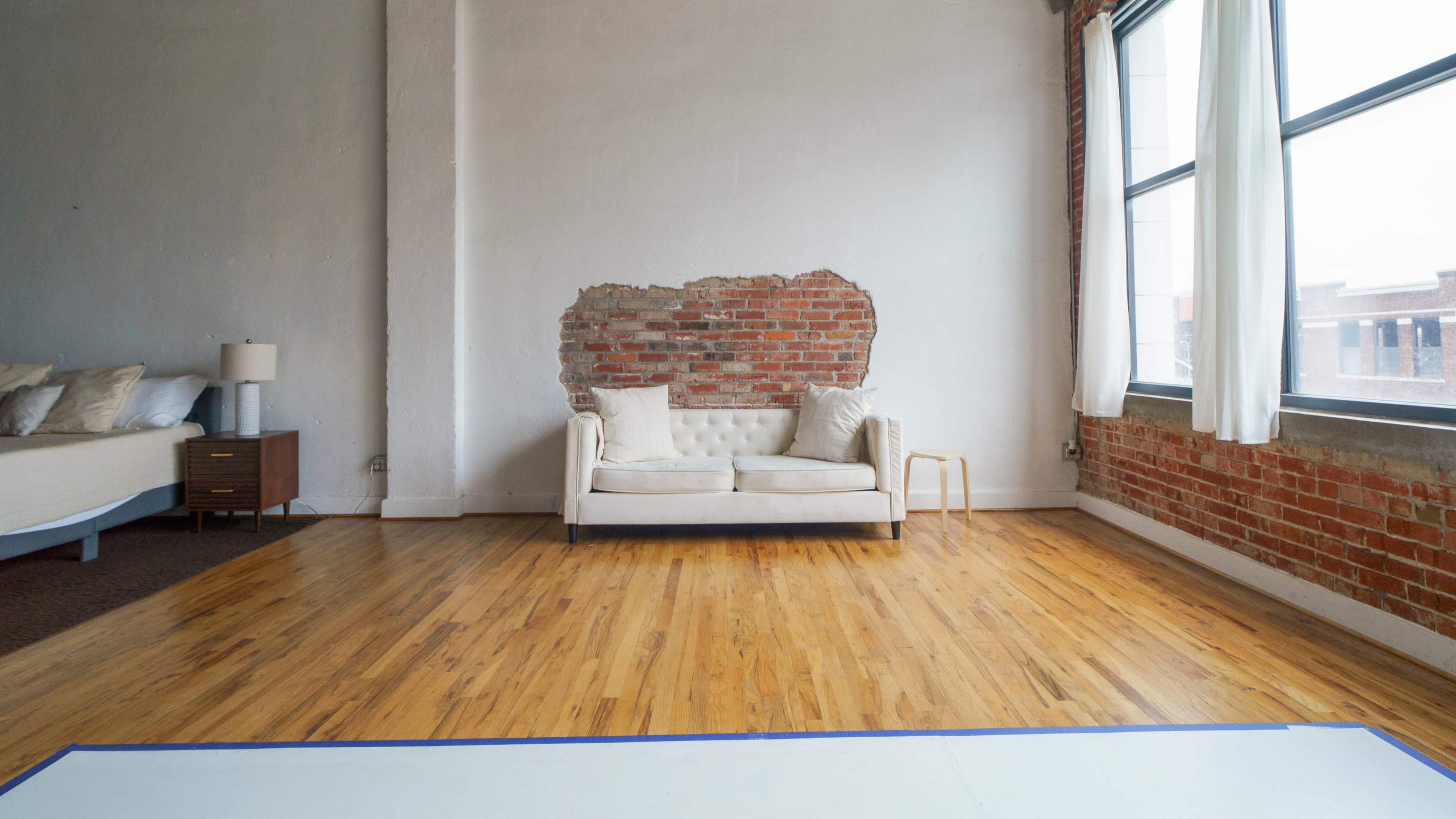 A minimalist room with exposed brick walls, a light-colored sofa, and large windows allowing natural light to enter.