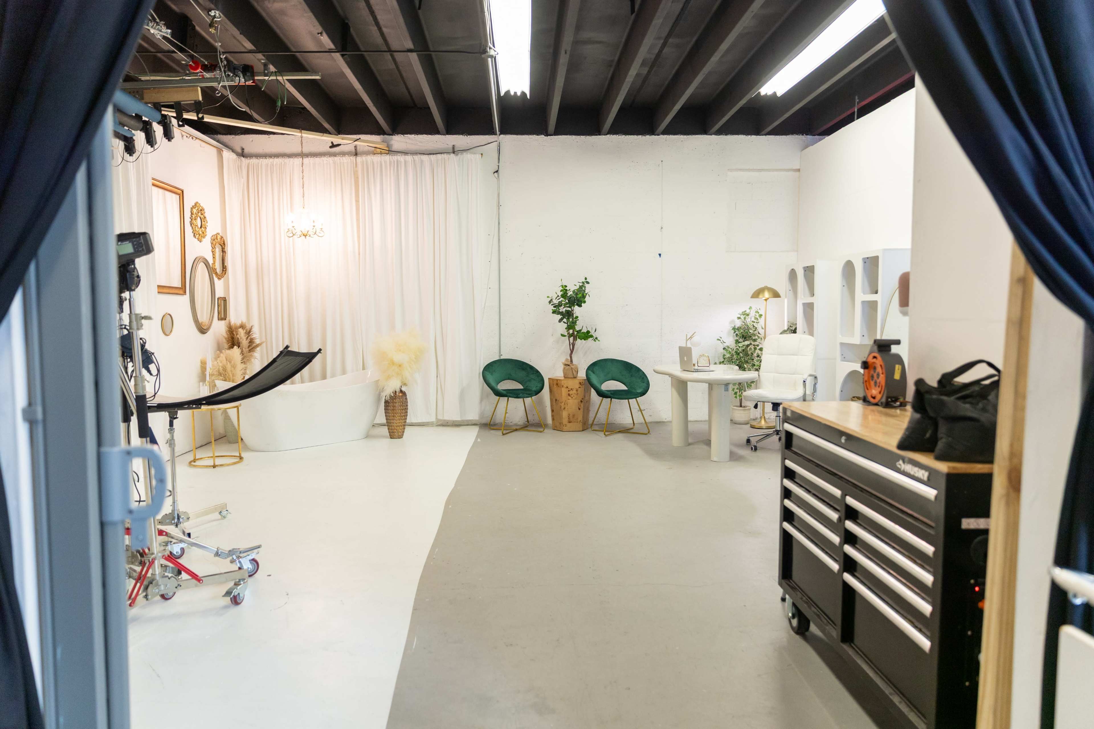 The image shows a well-lit studio space with a white backdrop, a bathtub, two green chairs, and a work table surrounded by decorative plants.