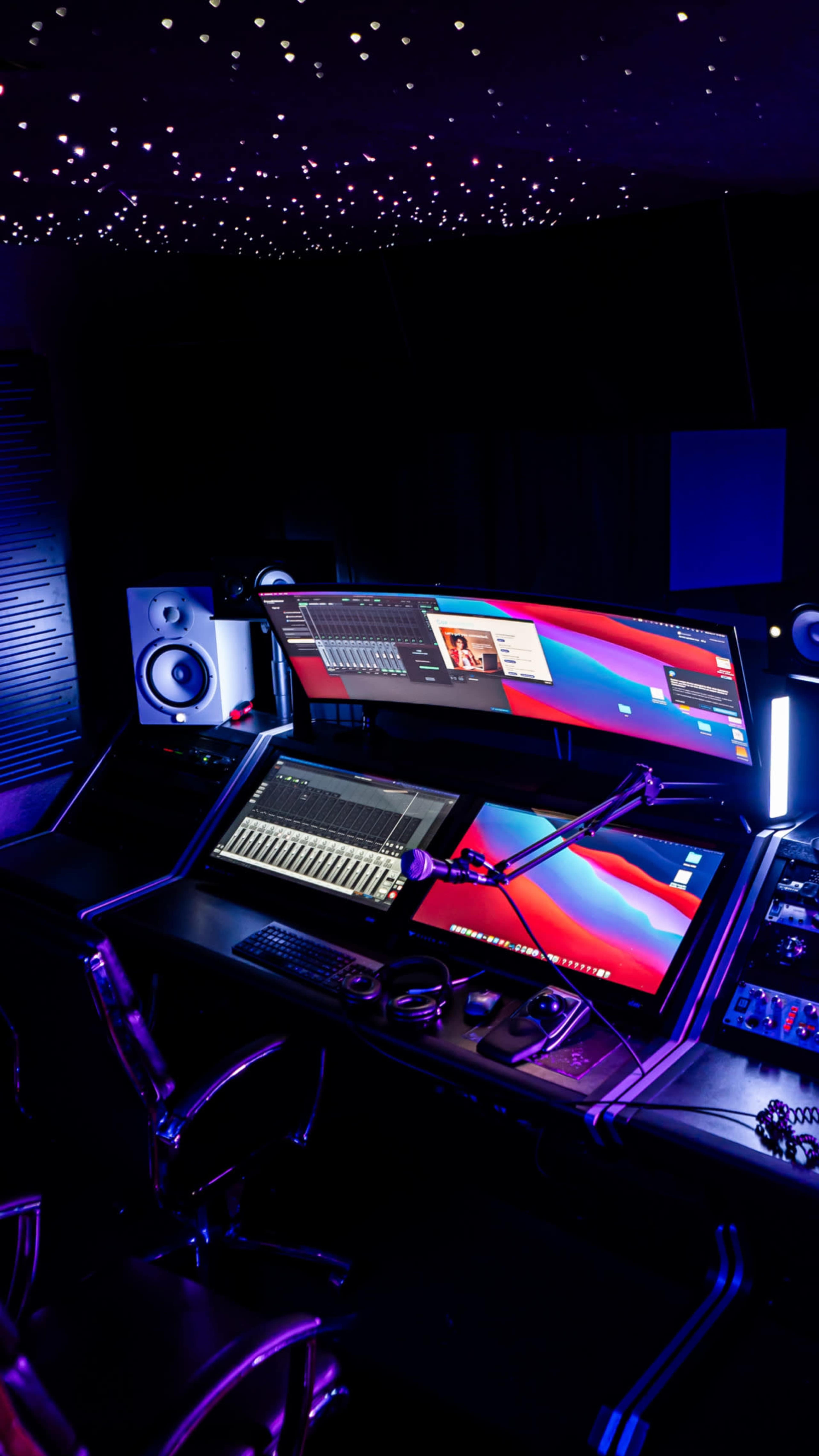A modern music production studio features dual computer monitors, a keyboard controller, and audio equipment illuminated by ambient lighting.