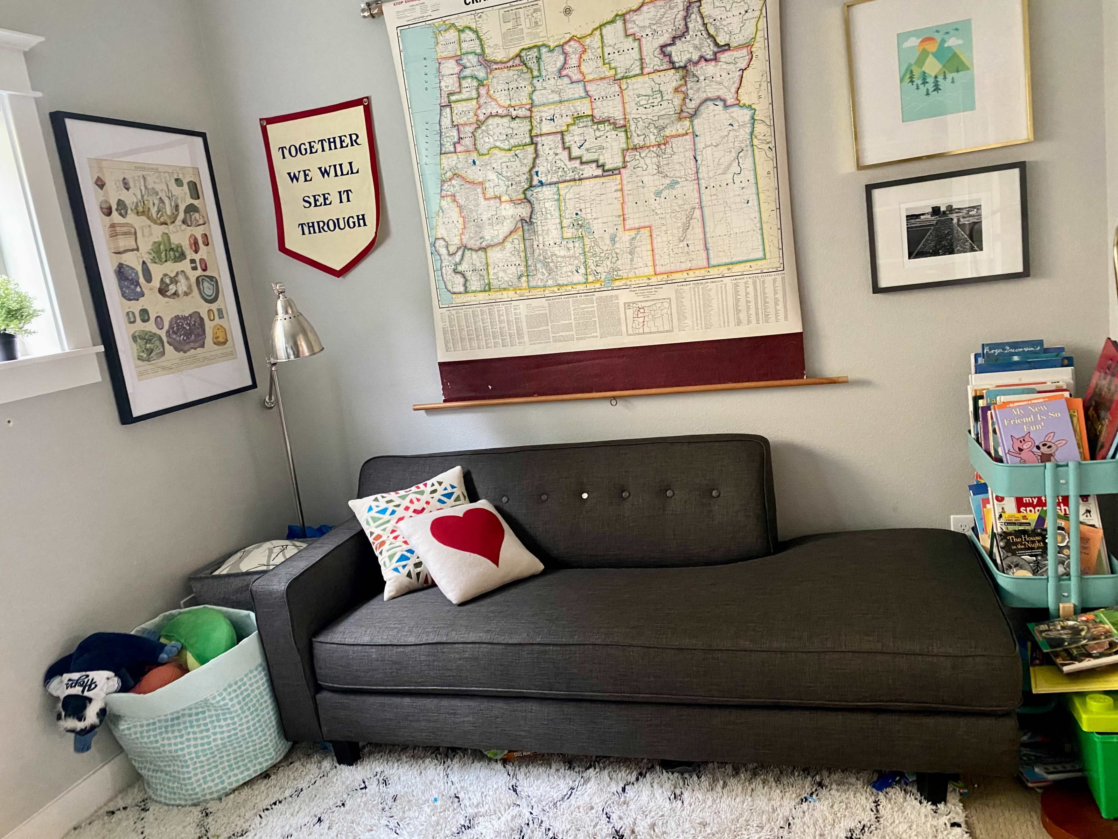 A gray chaise lounge with a red heart pillow is positioned against a wall adorned with maps and framed art, alongside shelves filled with colorful books and toys.