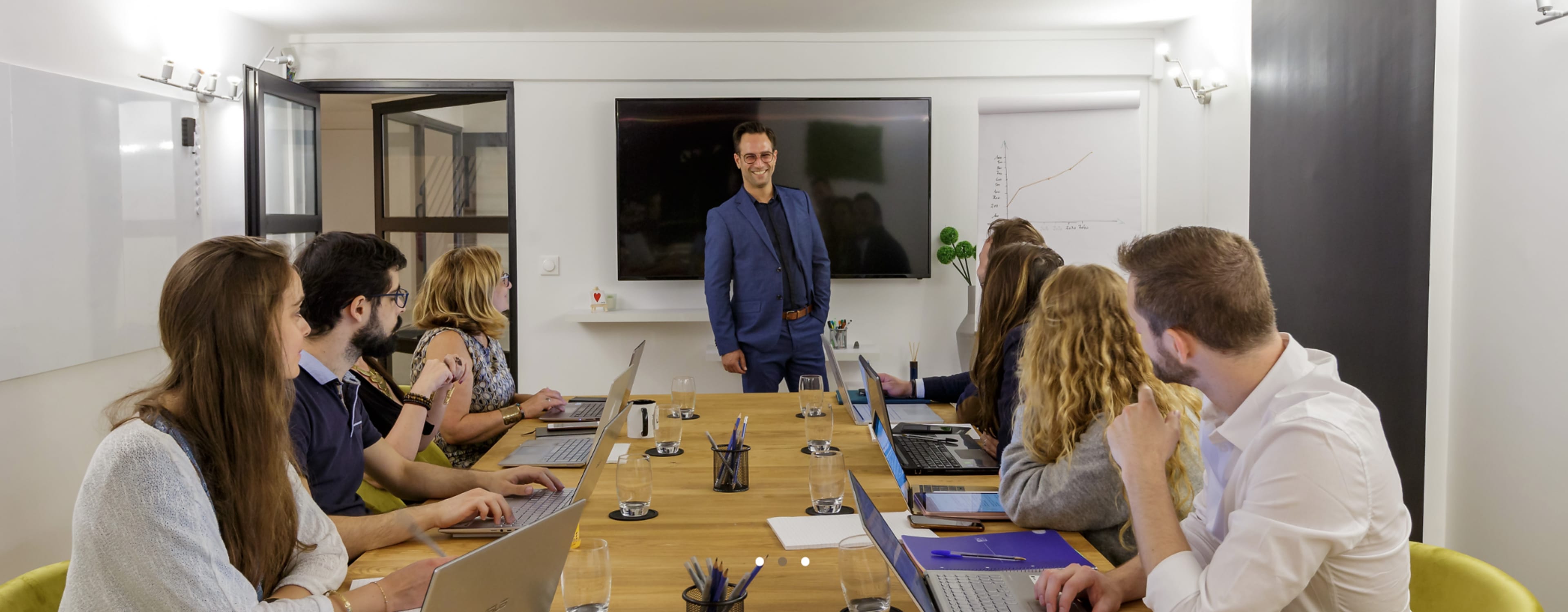 A group of six people with laptops sits at a table while a man in a blue suit presents in front of a screen in a conference room.