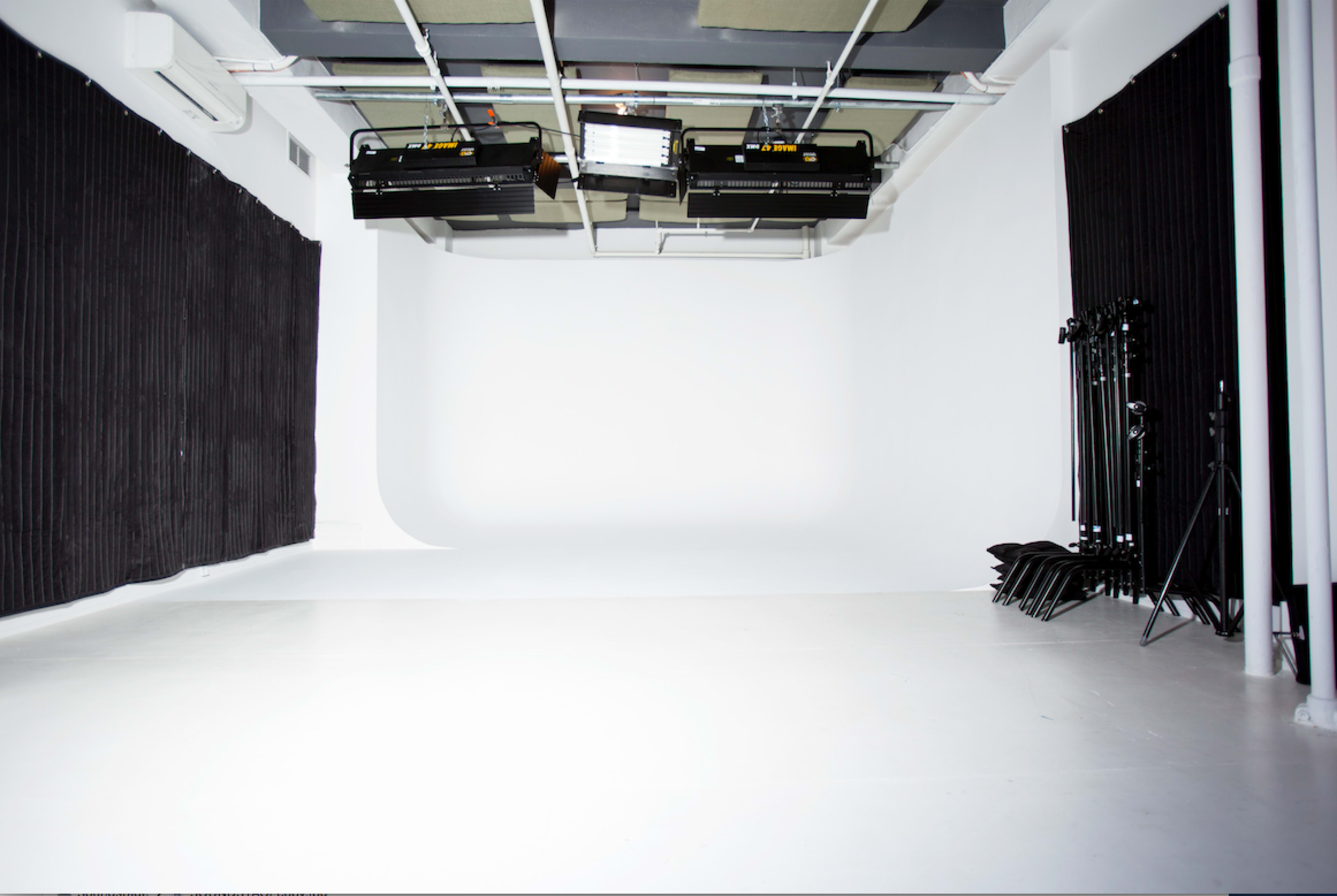 A photography studio with a seamless white backdrop, black curtains, overhead lights, and equipment stored at the side.