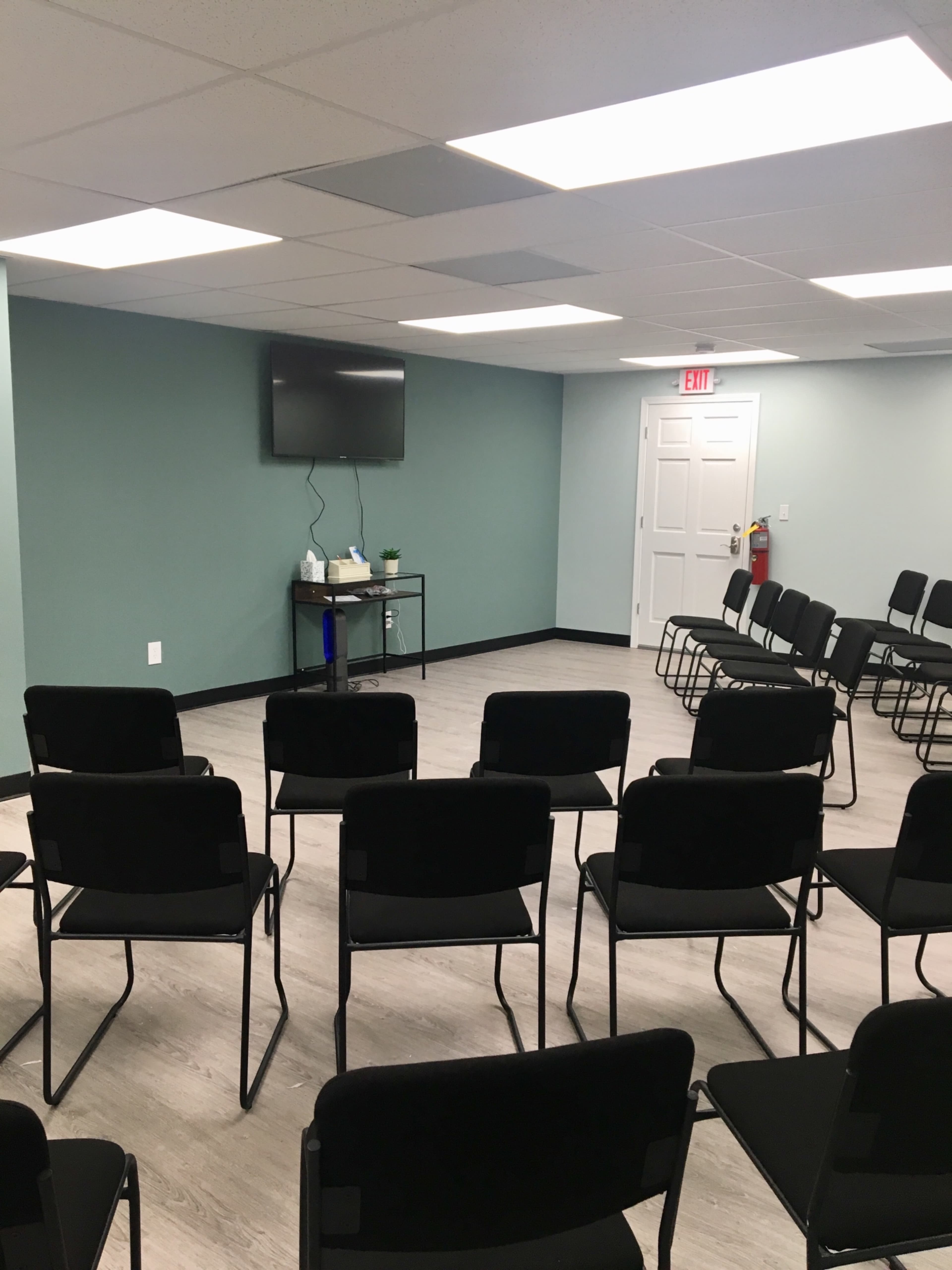 A meeting room with rows of black chairs facing a wall-mounted television, featuring light green walls and a door at the back.