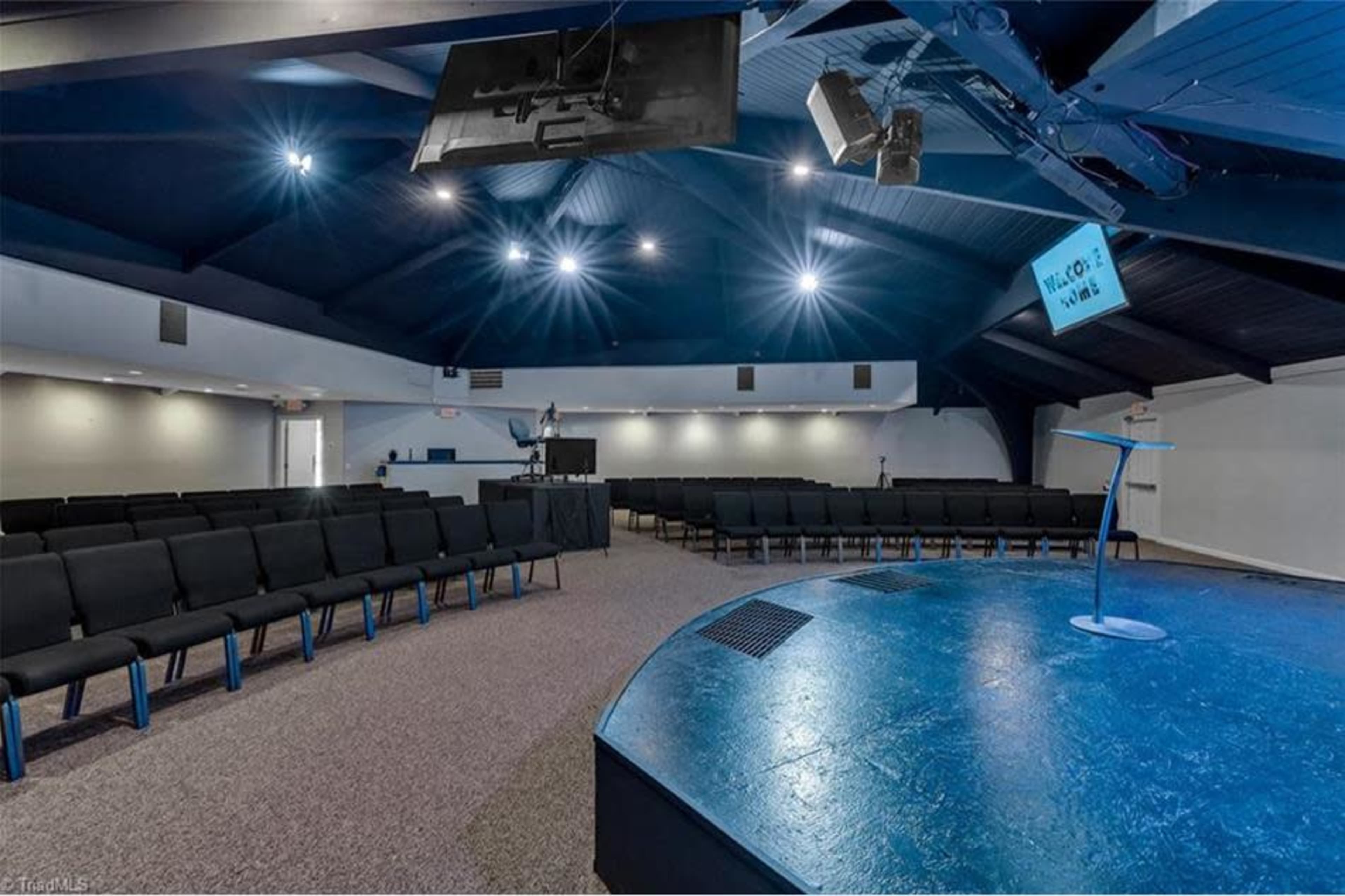 An empty auditorium with rows of black chairs facing a raised stage, featuring spotlights and a large screen on the wall.