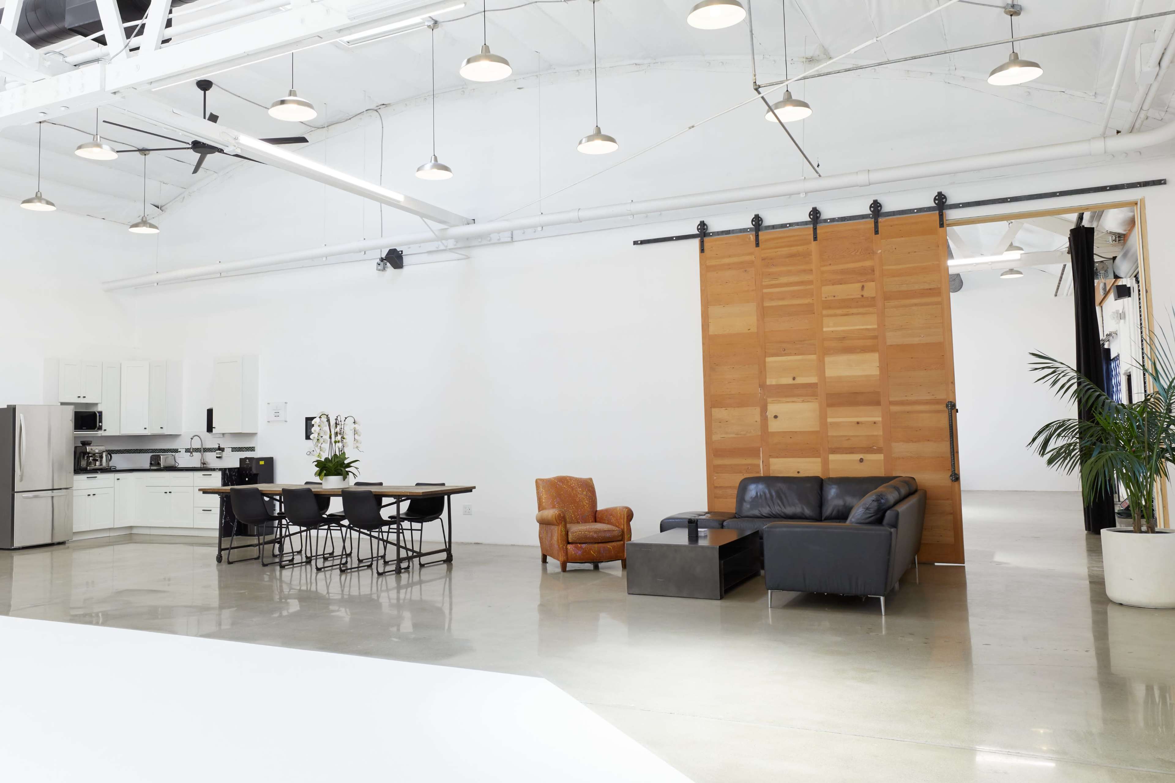The image shows a spacious, modern interior featuring a kitchen area with white cabinets, a dining table, and a living space including a leather sofa and an armchair, separated by a wooden sliding door.
