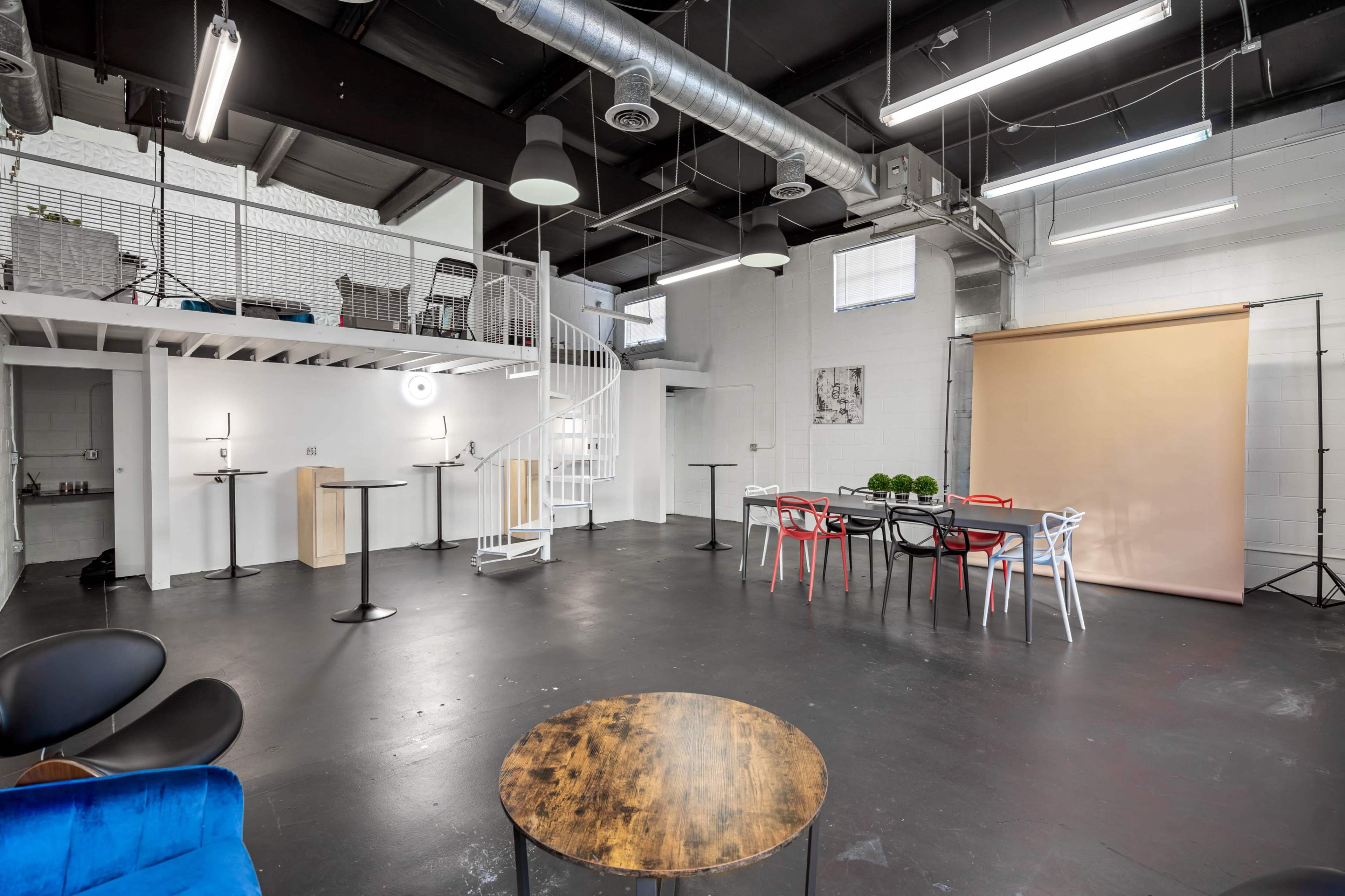 The image shows an industrial-style studio space featuring a spiral staircase, tables, chairs, and a backdrop for photography.