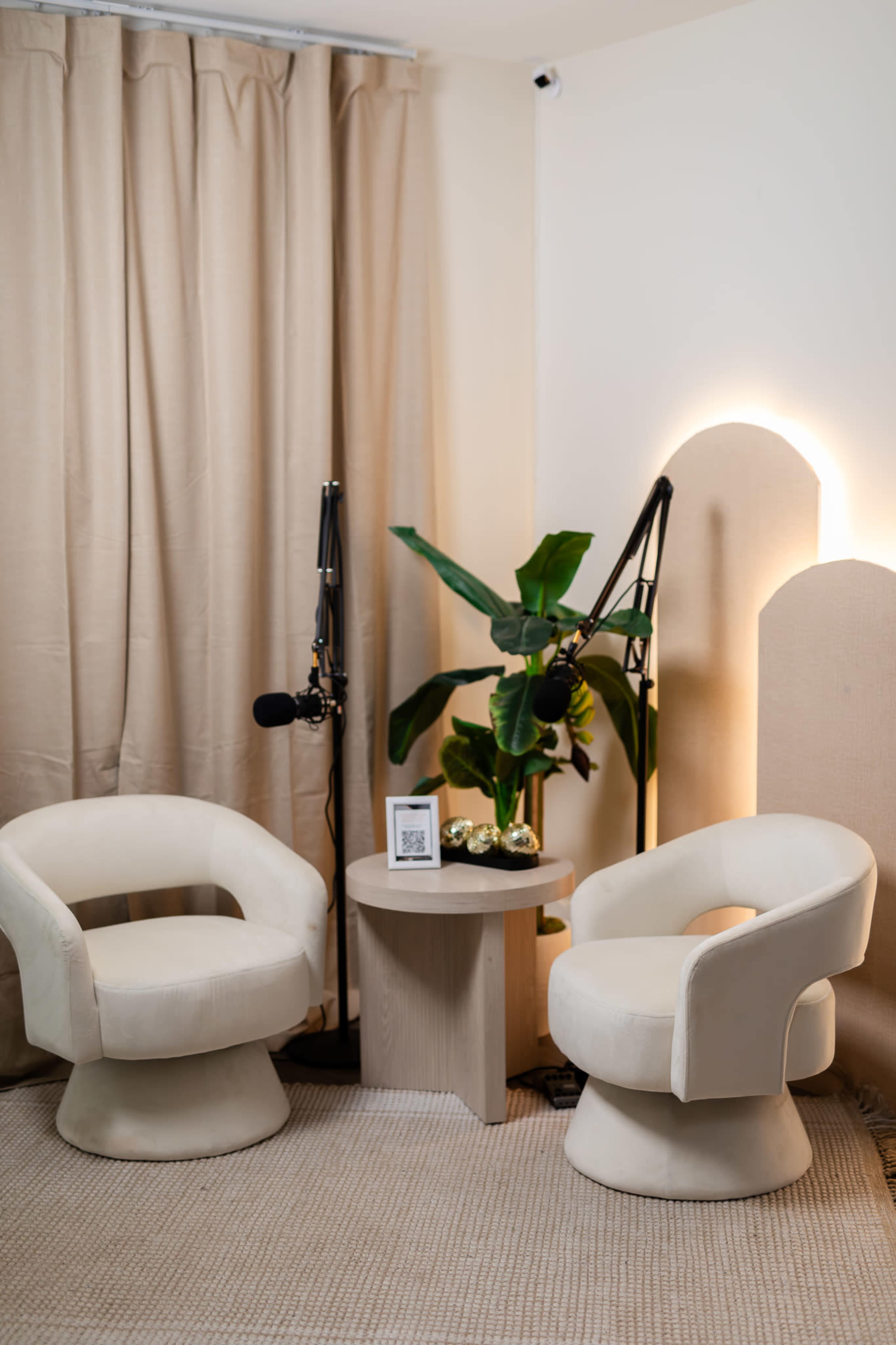 The image shows two sculptural white chairs facing a round wooden table, with a plant and microphones placed nearby against a backdrop of beige curtains.