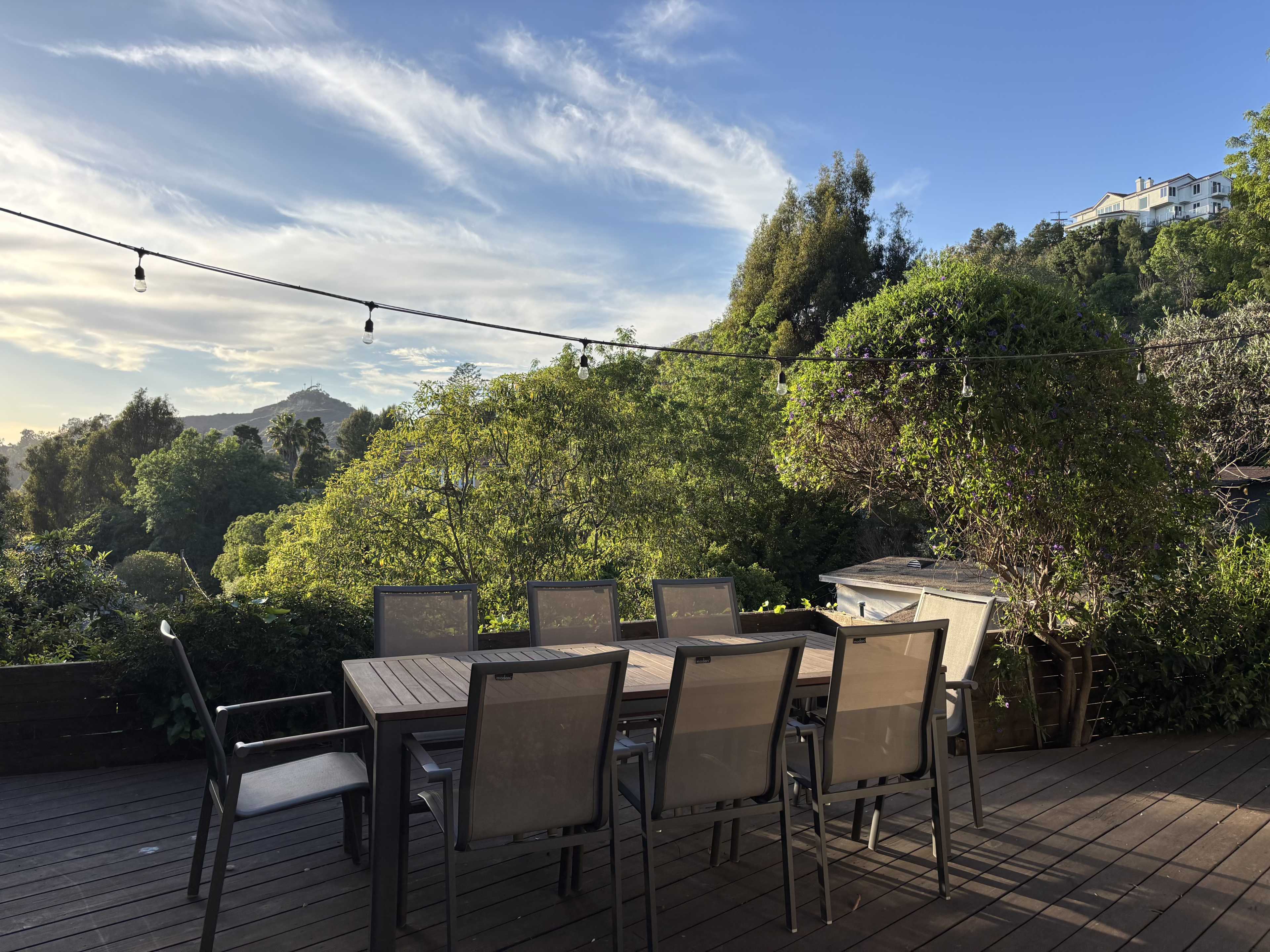 A wooden dining table with several chairs is set on a deck, overlooking lush greenery and hills under a clear sky.