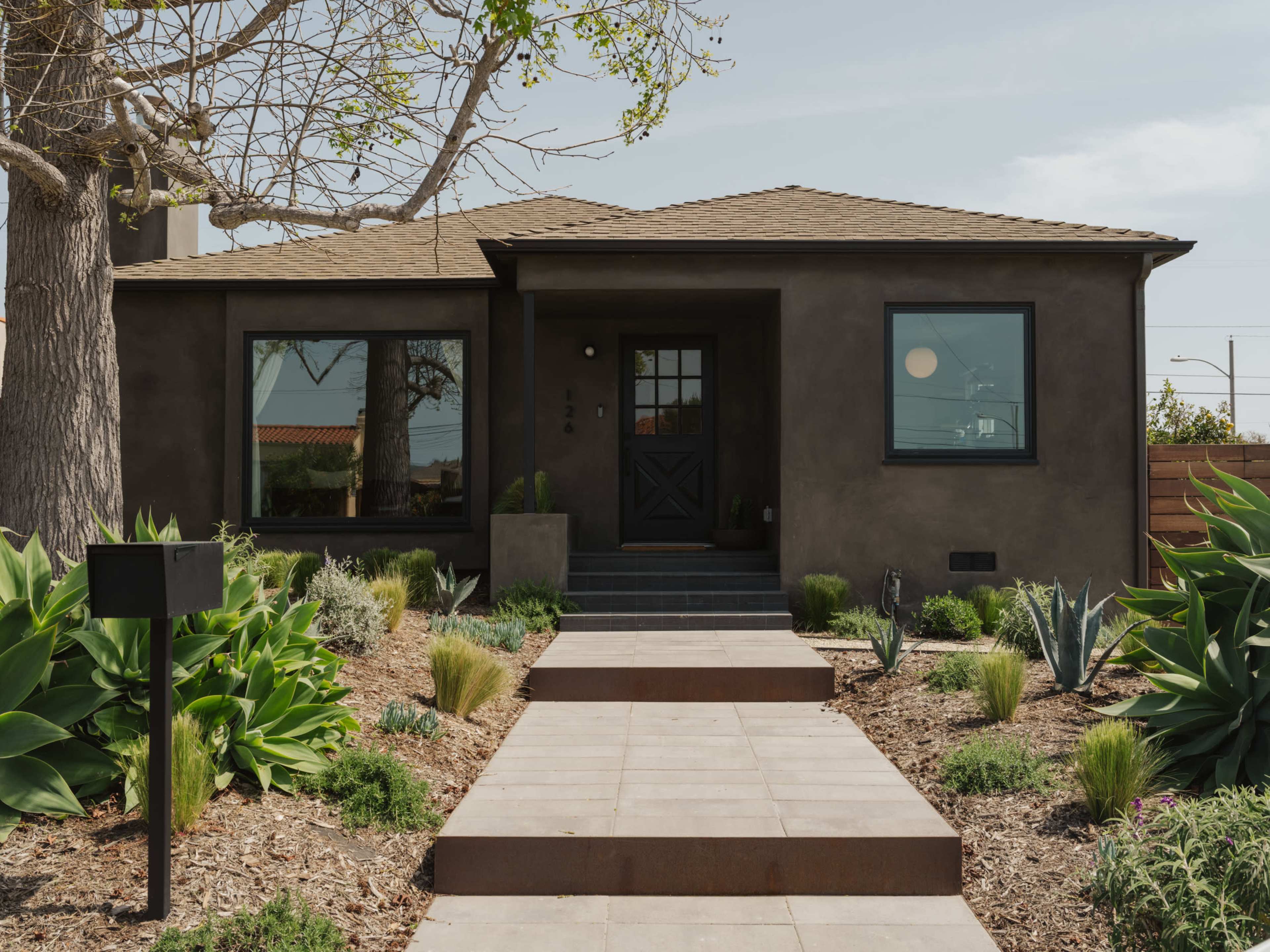 A single-story house with a brown stucco exterior and a simple pathway leads to the entrance, surrounded by landscaped greenery and desert plants.