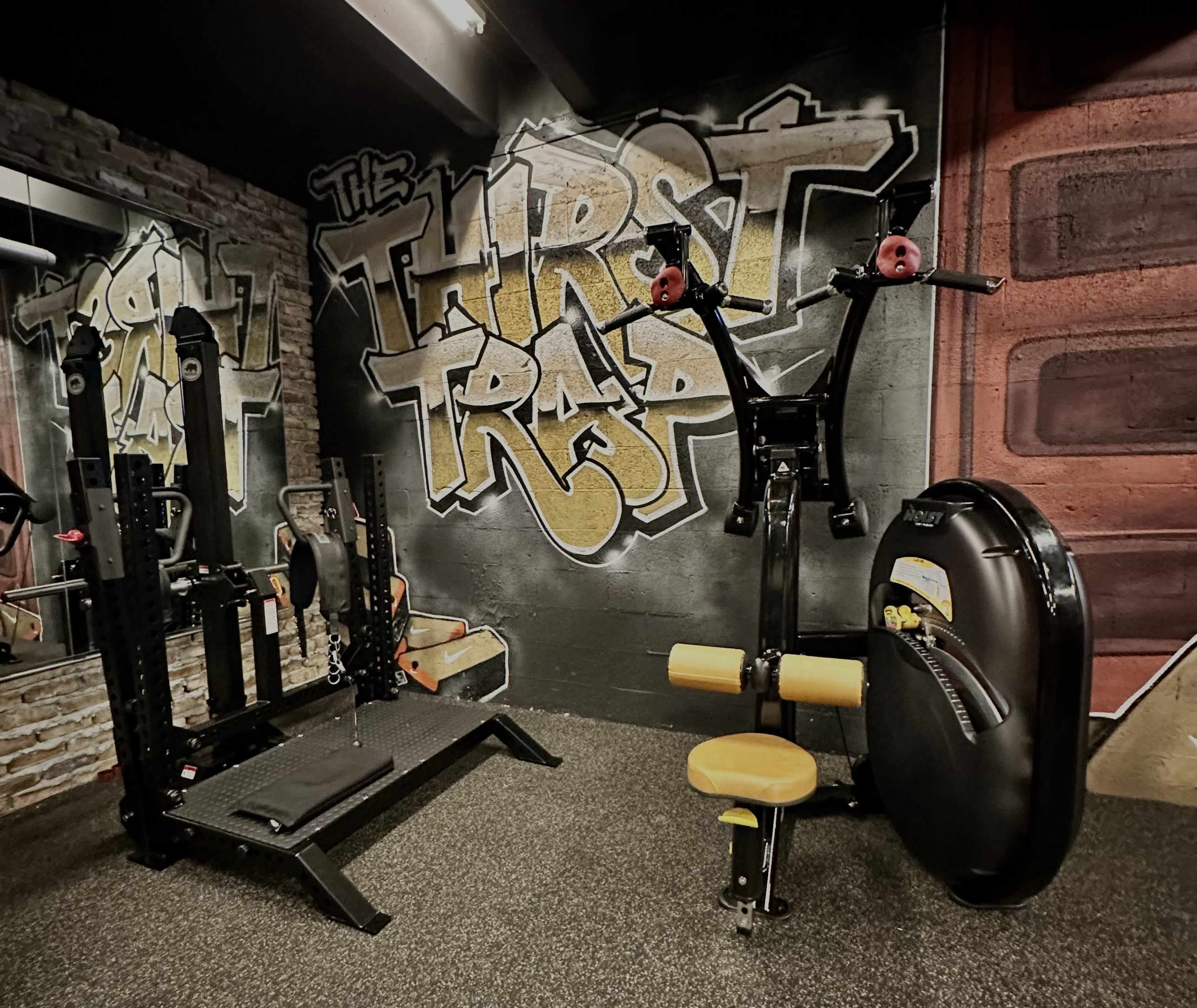 A gym interior featuring workout equipment against a graffiti-style wall mural that reads "The Thirst Trap."