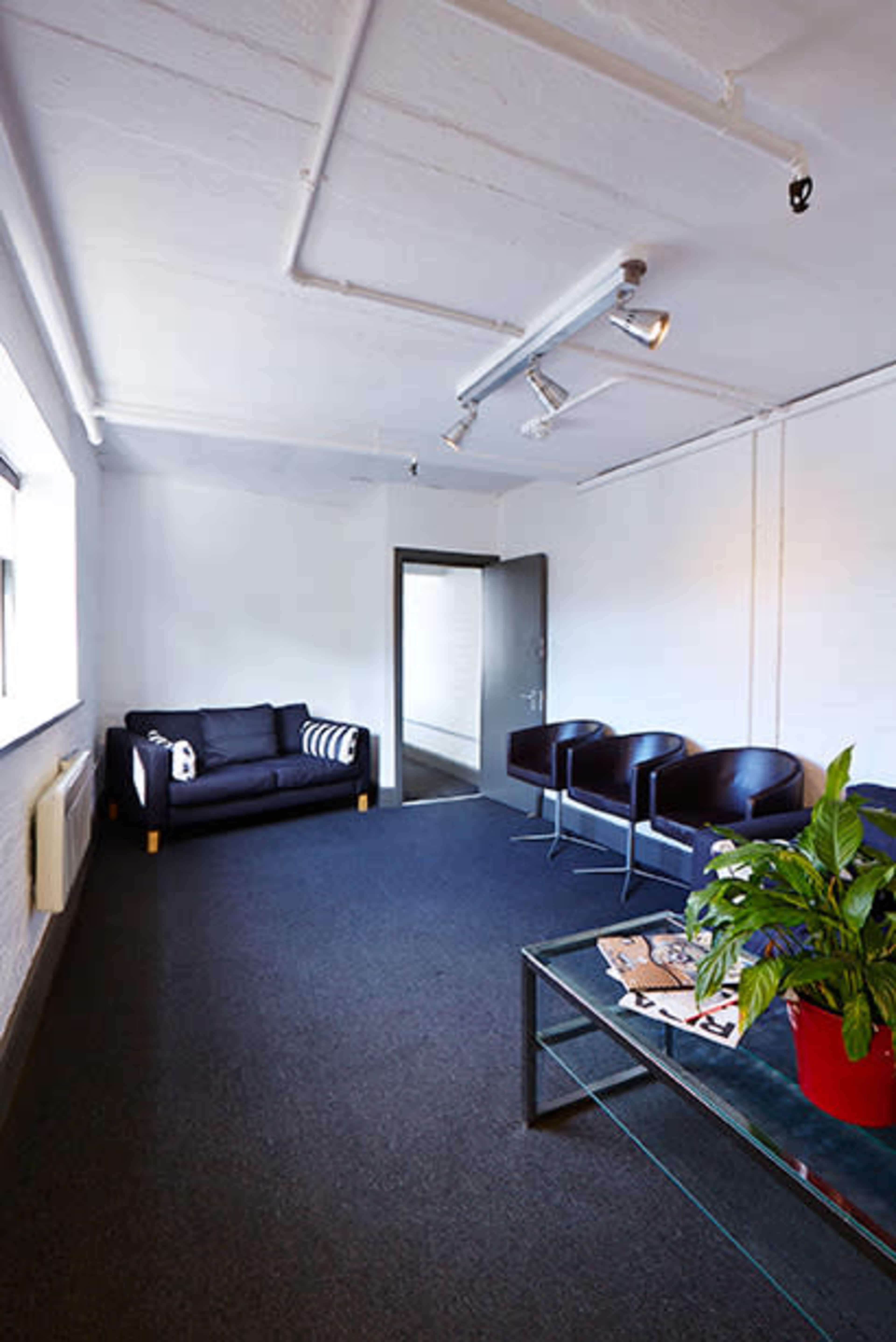 A minimalist office space with a black sofa, two black chairs, a coffee table, and a potted plant against a light-colored wall.