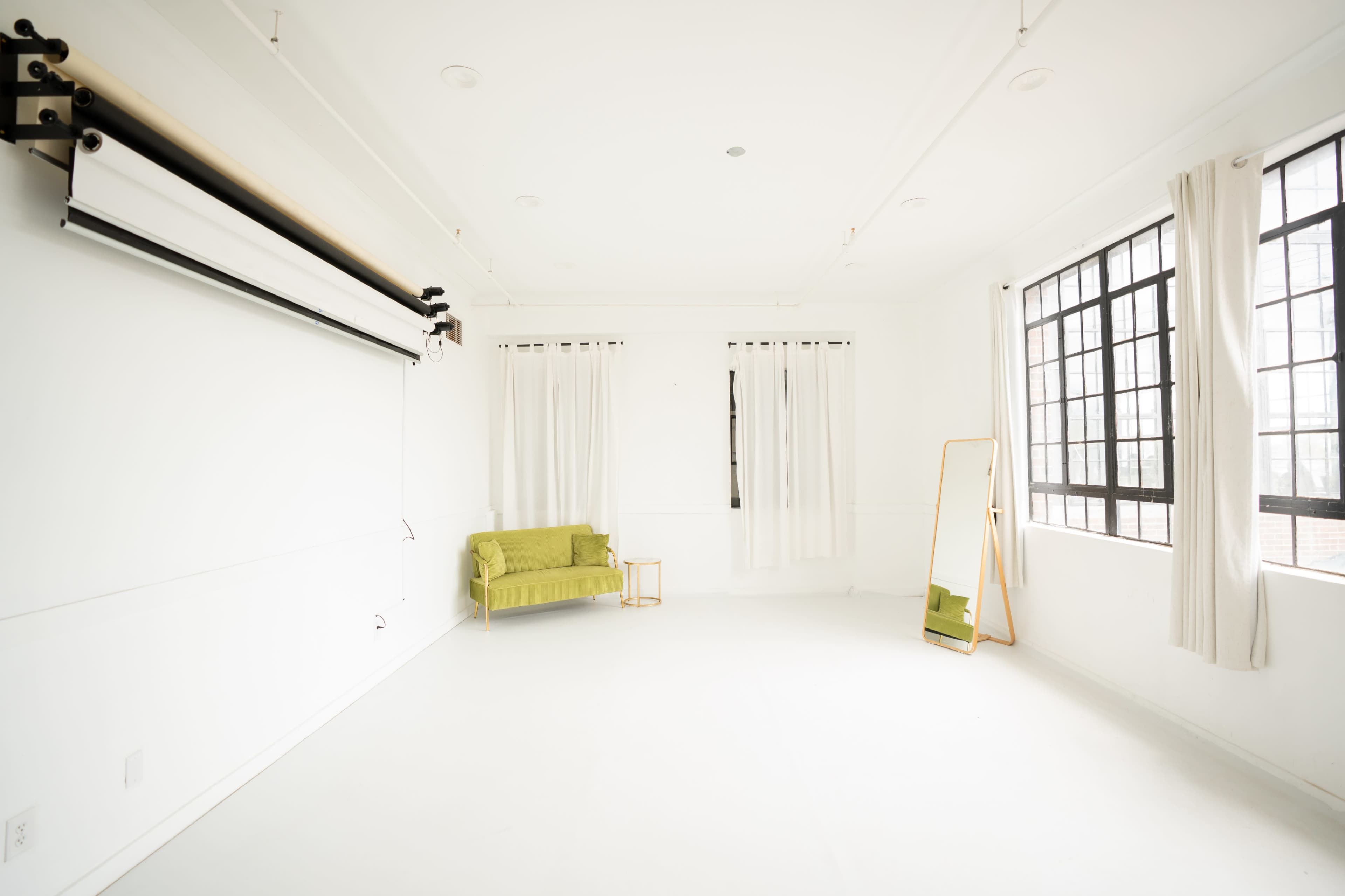 The image shows a minimalist room with white walls, a green sofa, a full-length mirror, and large windows with sheer curtains.