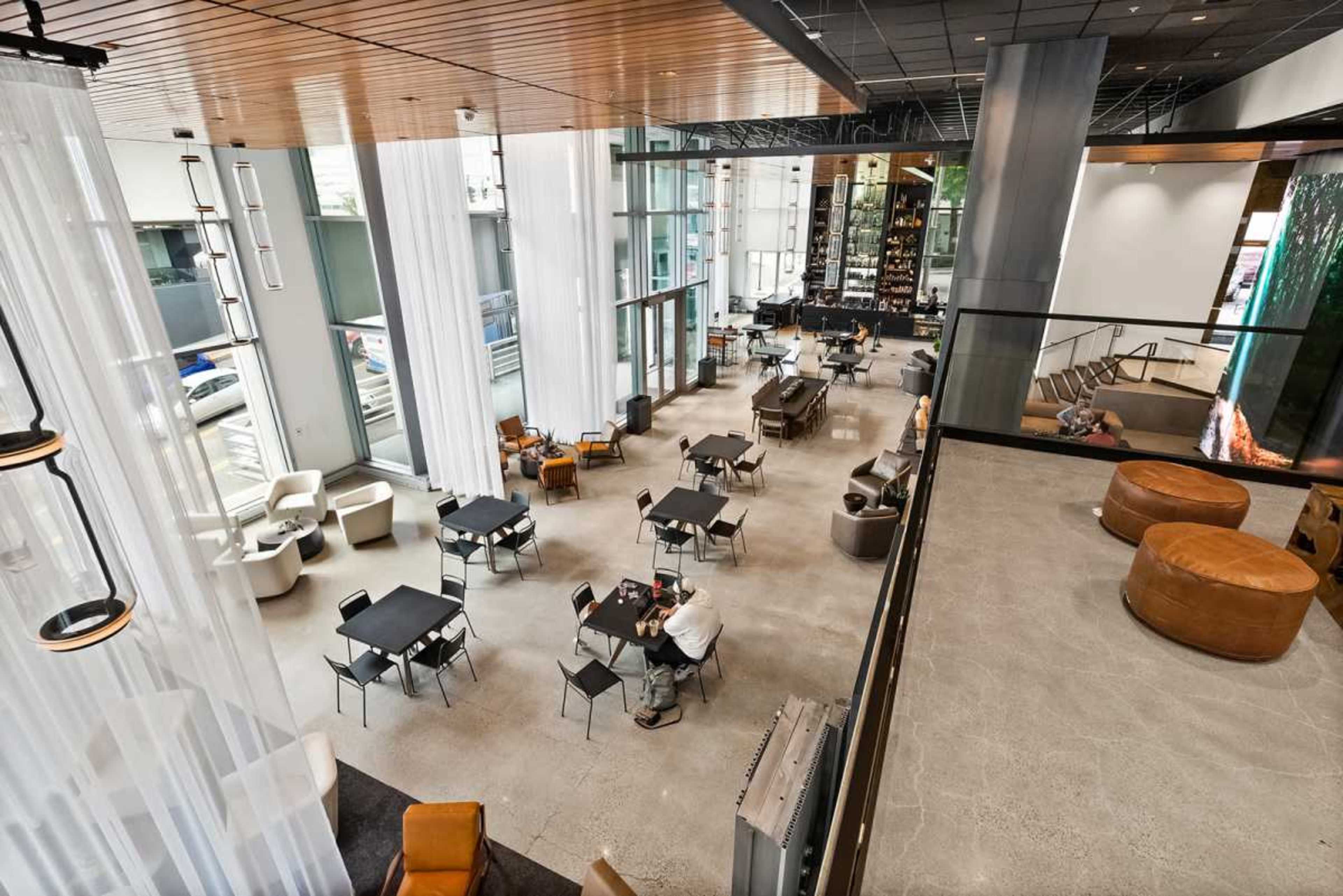 The image shows a large, modern lounge area with multiple seating arrangements, tables, and large windows allowing natural light to enter.