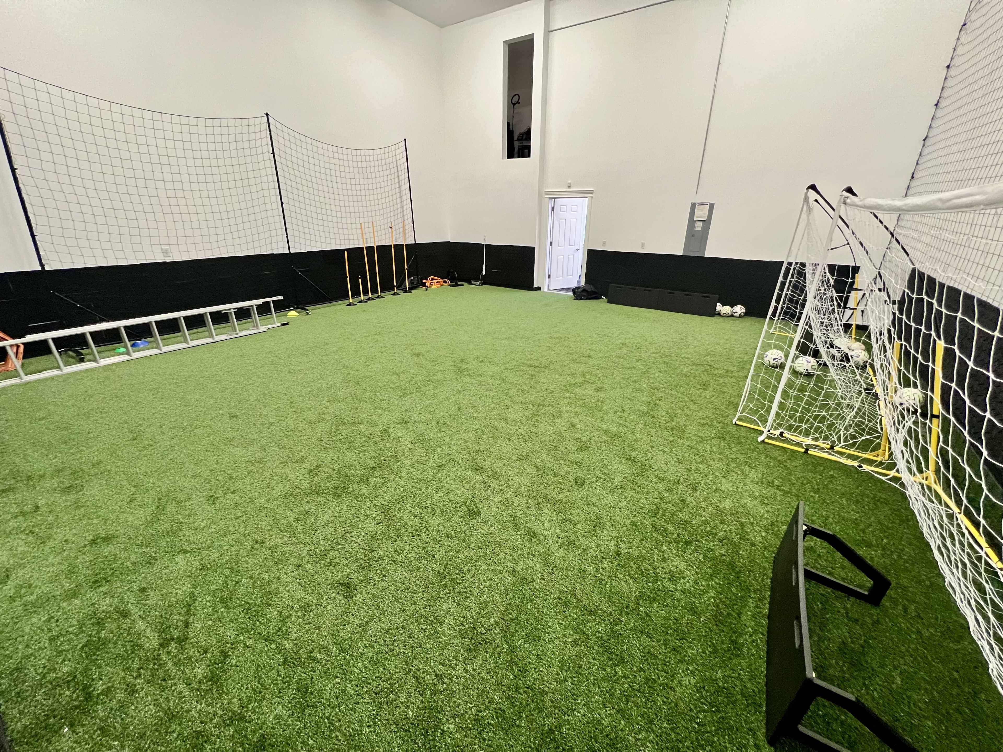 The image shows an indoor soccer training facility with artificial turf, two small goals, and training equipment along the walls.