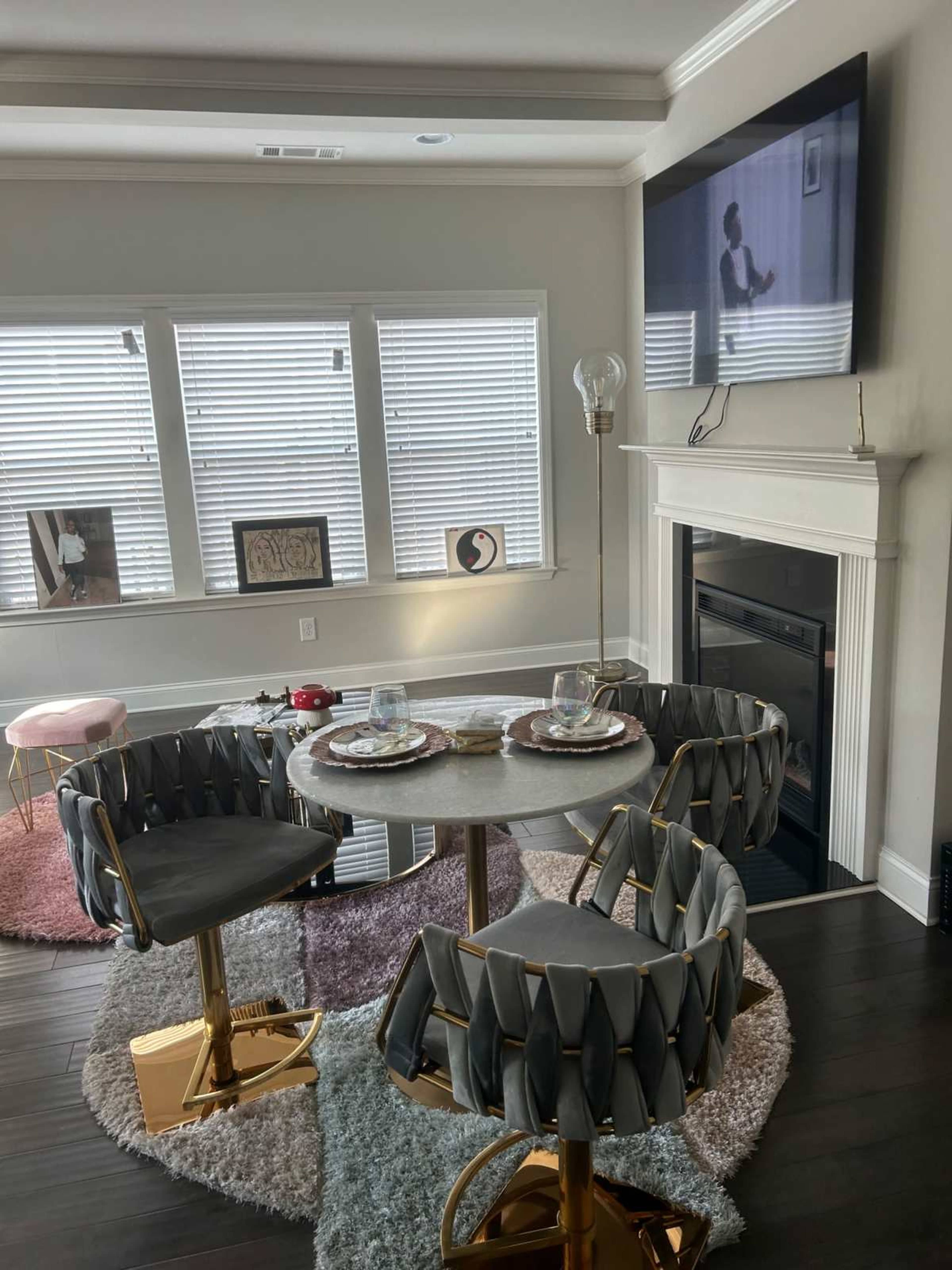 A modern dining area features a round table with two designer chairs, set with glassware and plates, alongside a fireplace and a television.