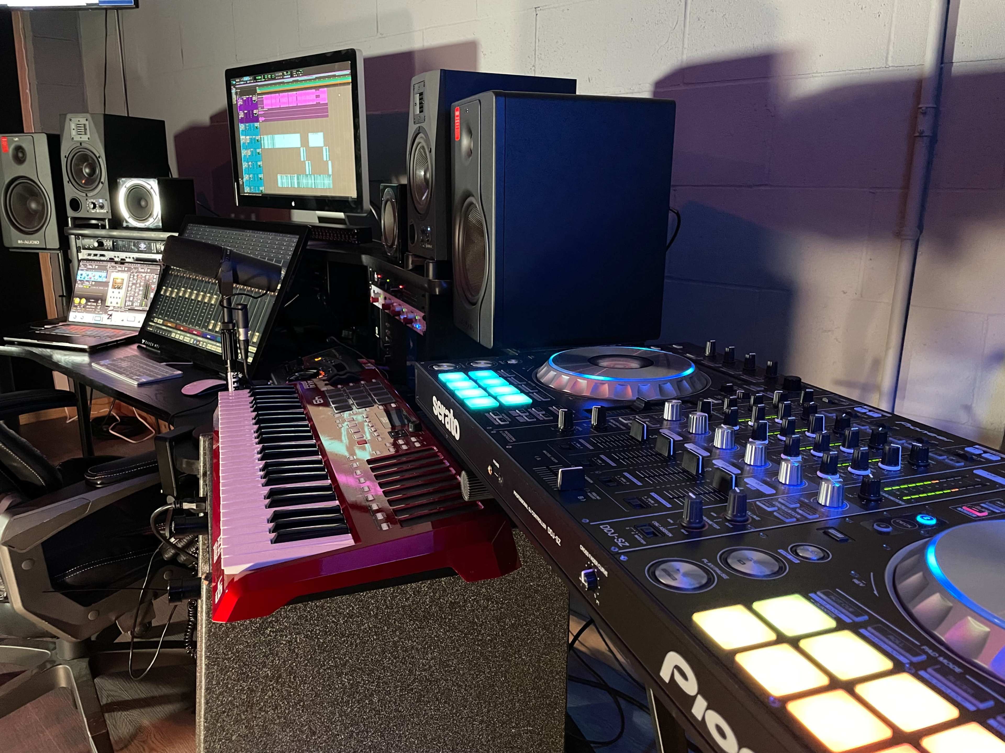 The image shows a modern music production studio with a keyboard, DJ controller, audio monitors, and a computer display showcasing a music editing software interface.