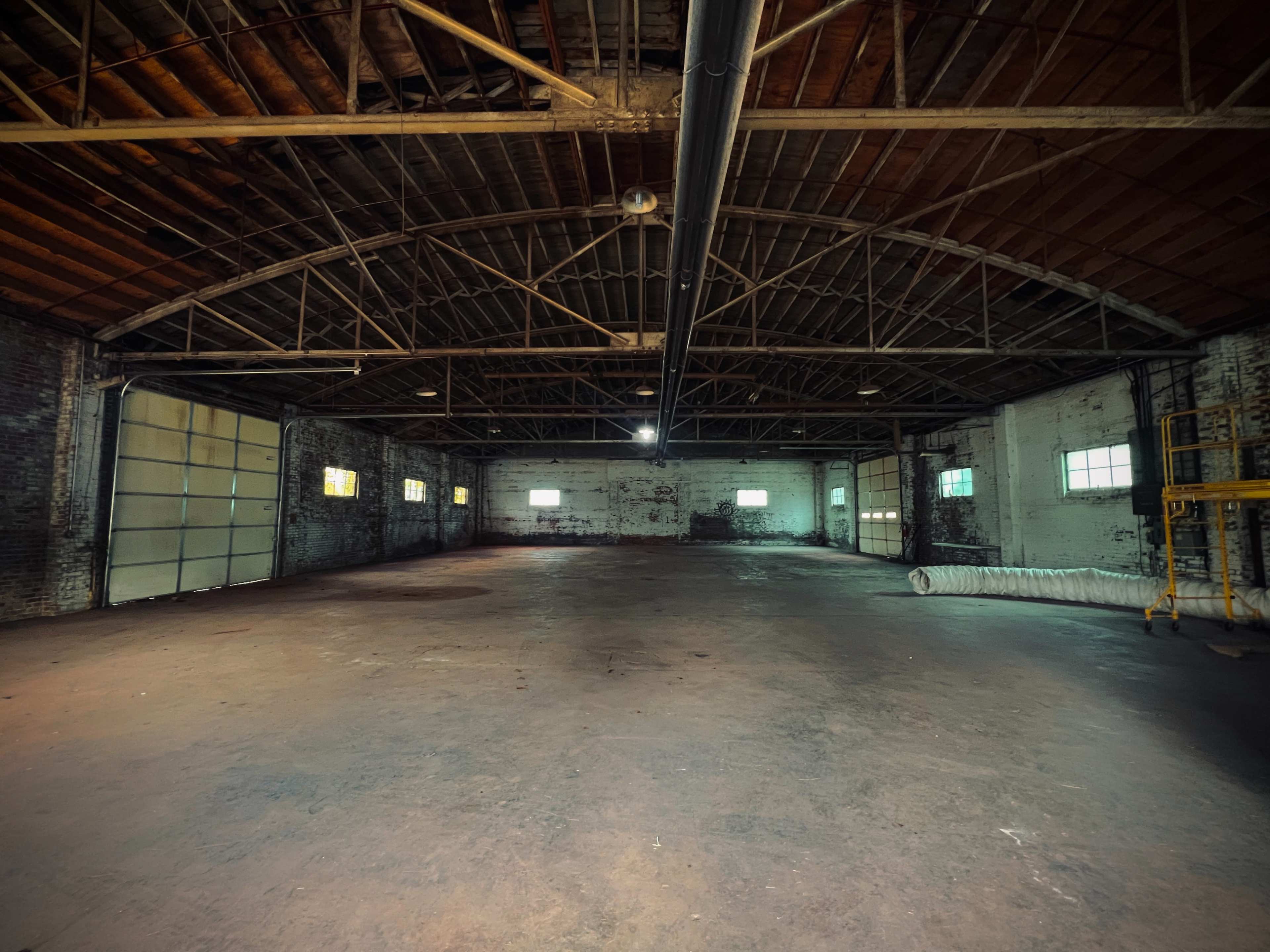 The image shows an empty industrial warehouse with exposed beams, concrete flooring, and multiple windows allowing light to enter.
