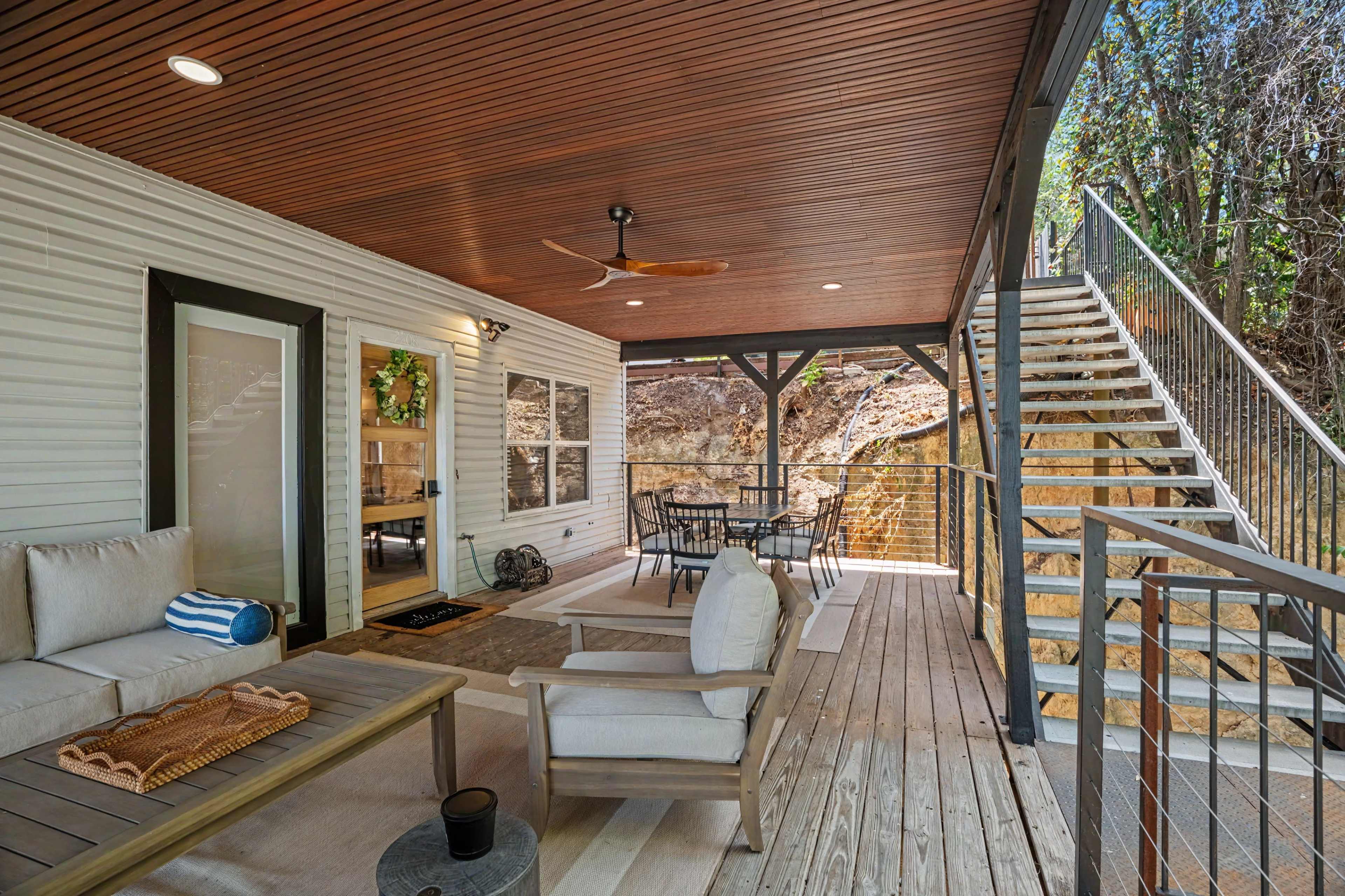 The image shows a covered outdoor patio area with seating, a coffee table, and a staircase leading to an upper level.