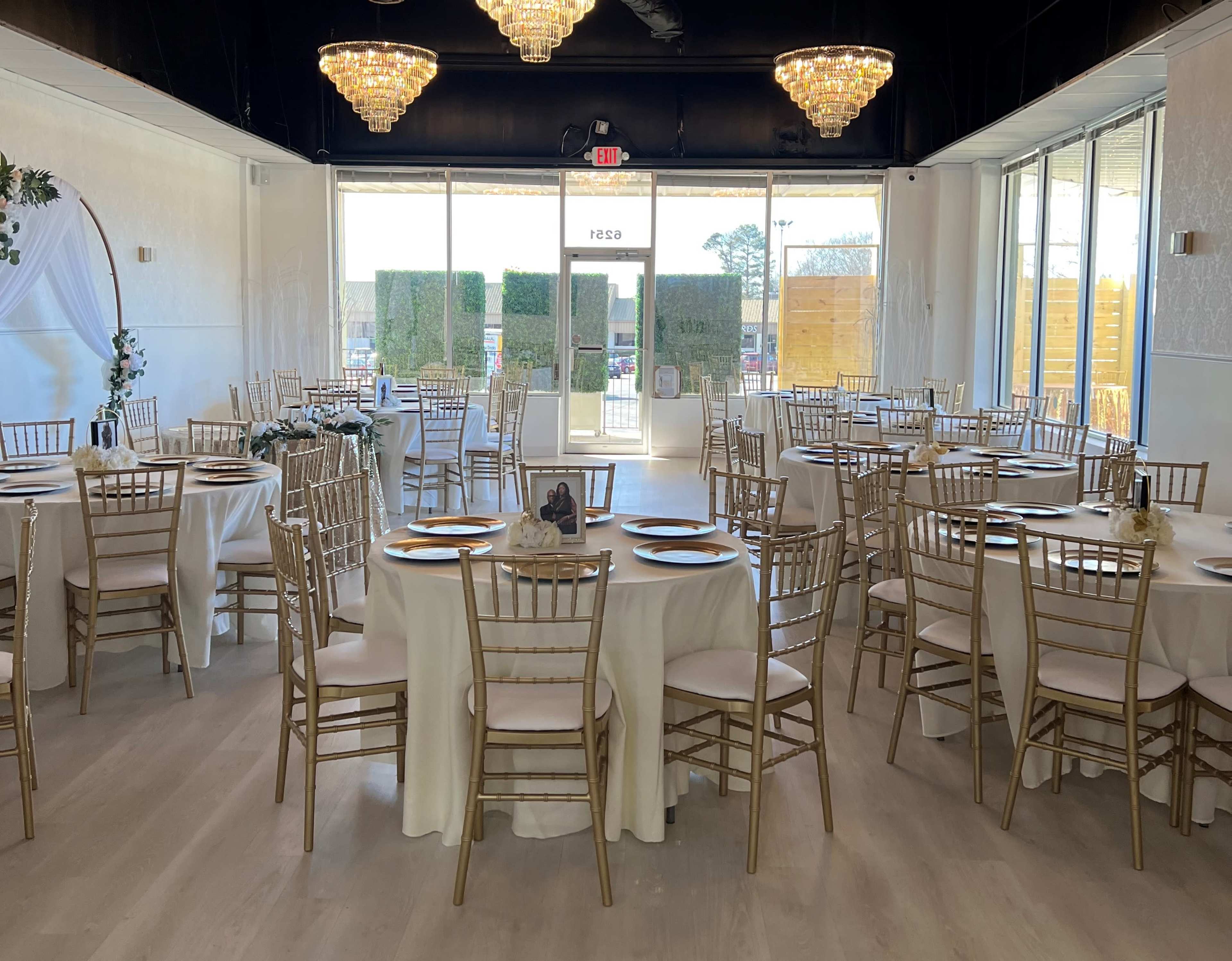 A dining hall set with round tables covered in white tablecloths and adorned with golden chairs, illuminated by chandeliers overhead.