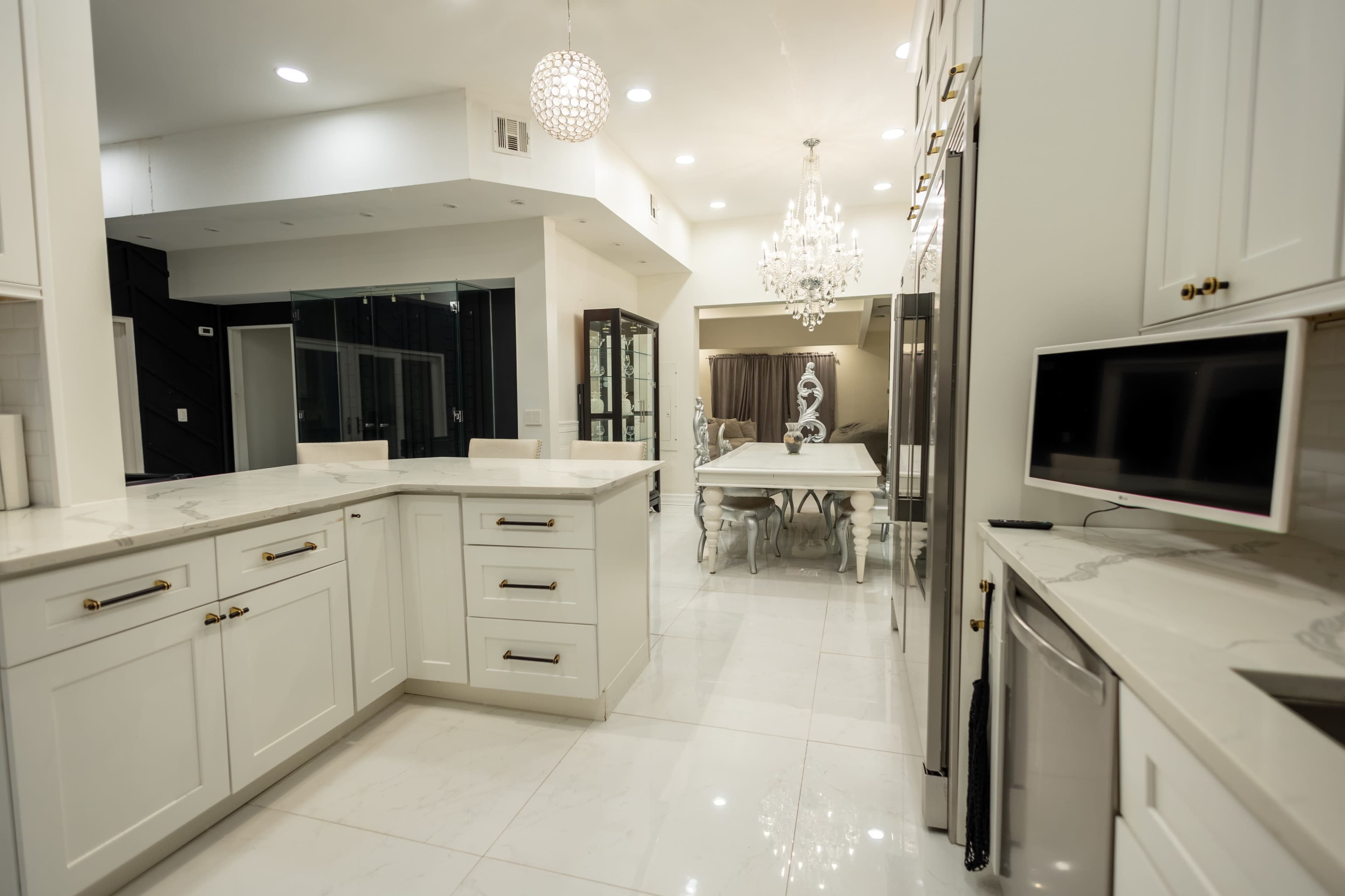 The image depicts a modern kitchen with white cabinetry and sleek countertops, leading into an elegant dining area with a chandelier and a glass table.