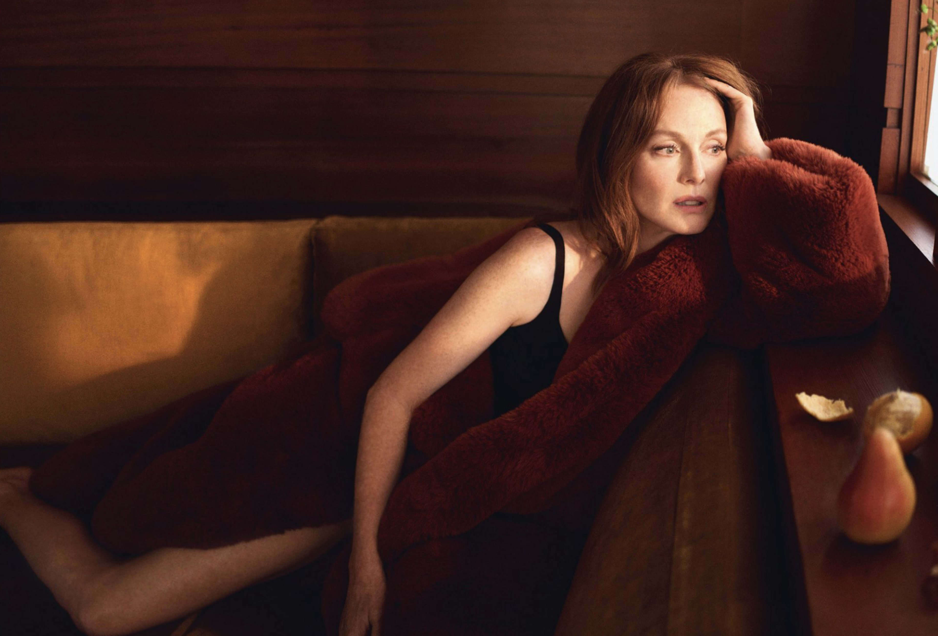 A woman with red hair rests on a couch, draped in a thick blanket, with a contemplative expression.