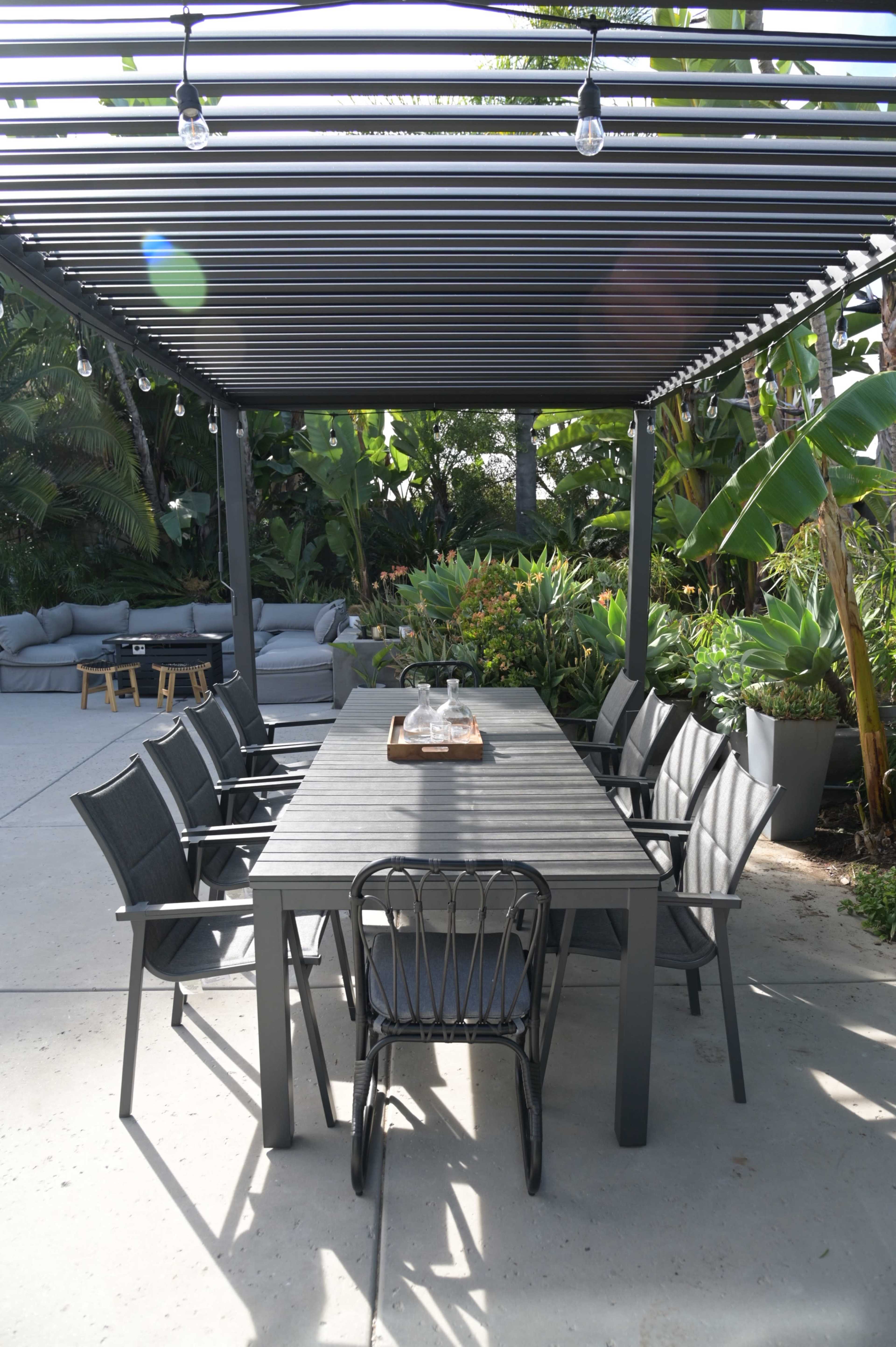A large outdoor dining table with multiple chairs is situated under a slatted pergola surrounded by lush greenery.