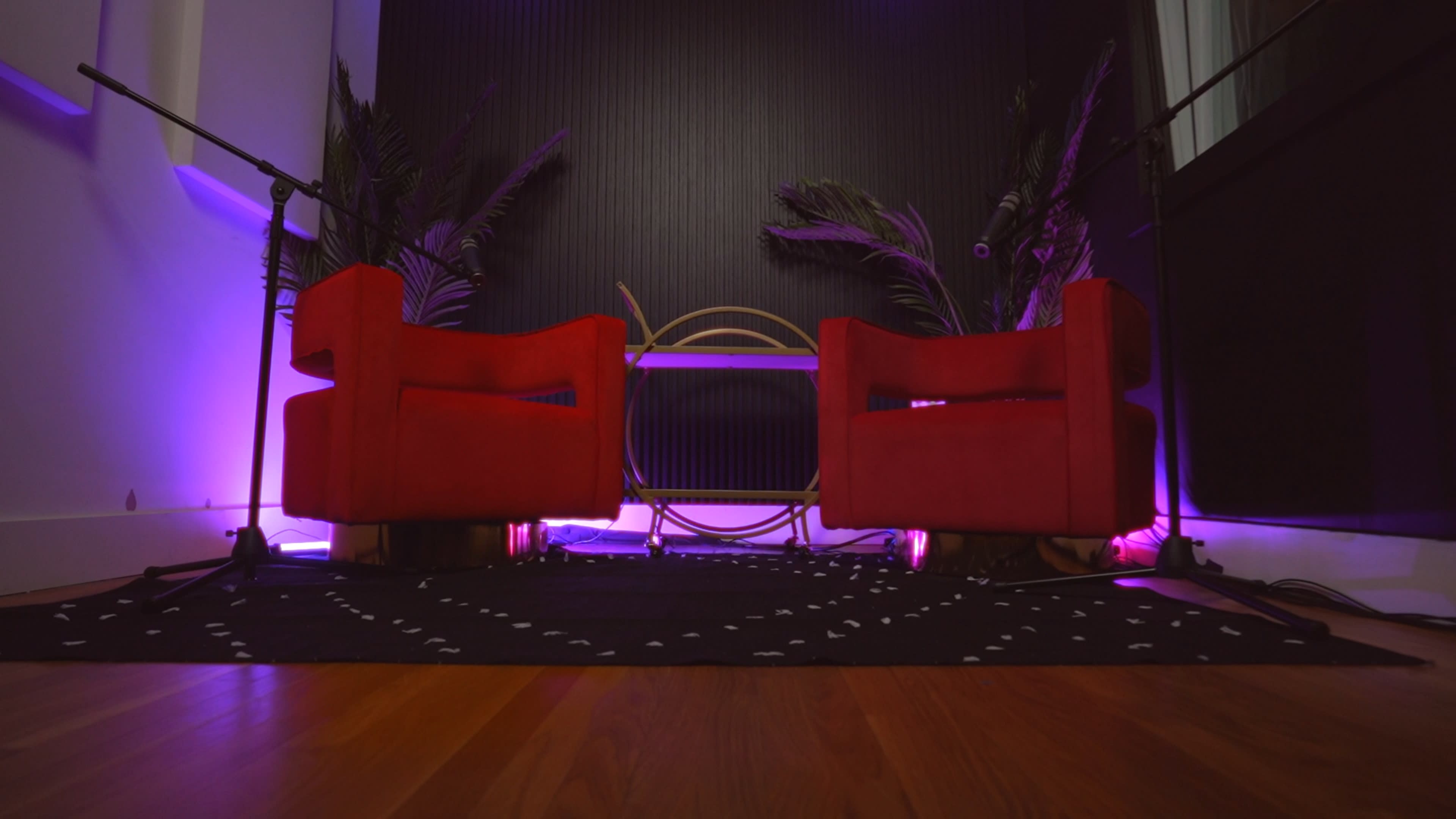 The image features two red chairs facing each other across a circular gold table, surrounded by plants and illuminated by purple lighting against a dark wall.