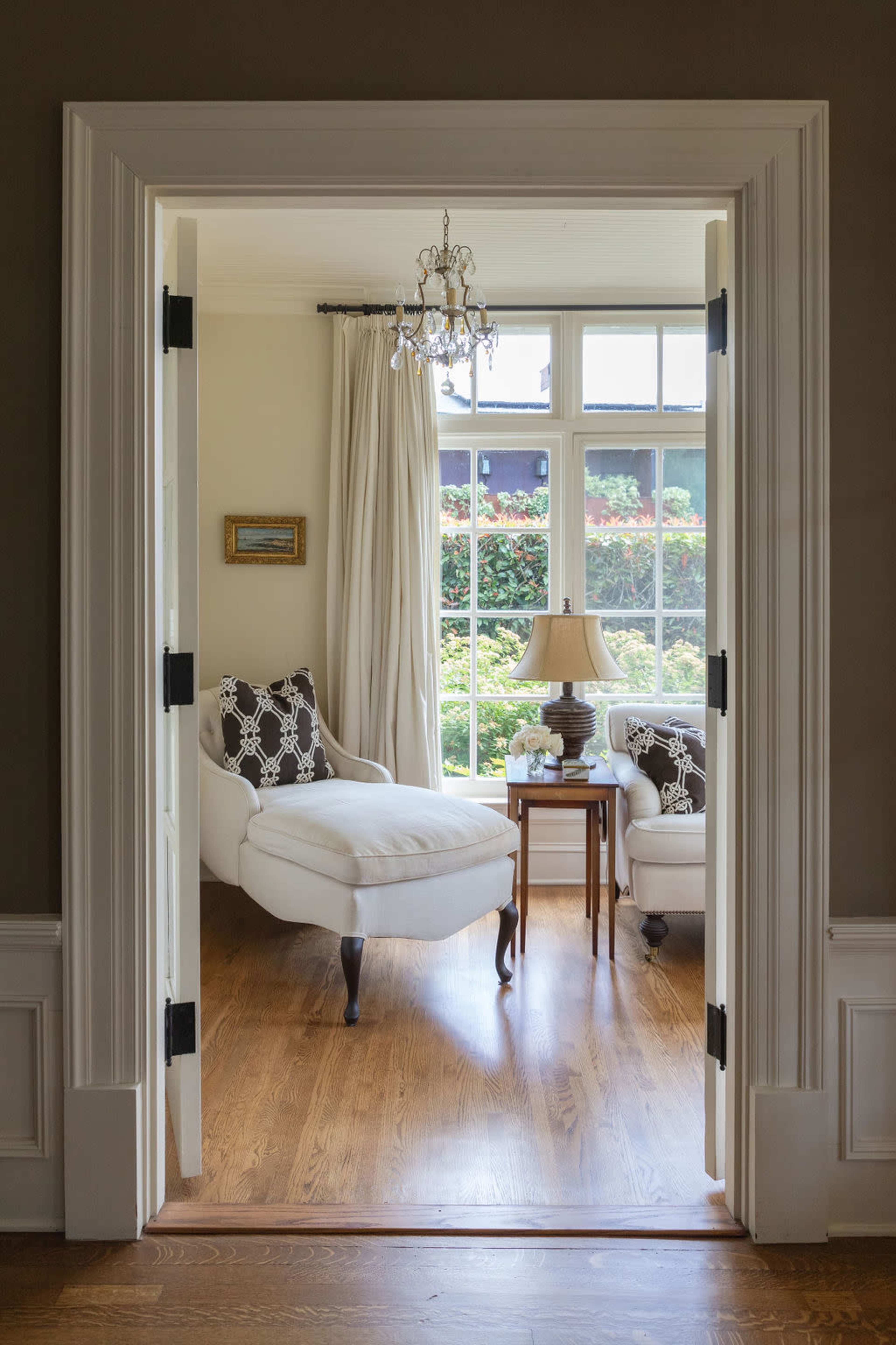 A well-lit living room with a chaise lounge and a side table beside a window, framed by an open doorway.