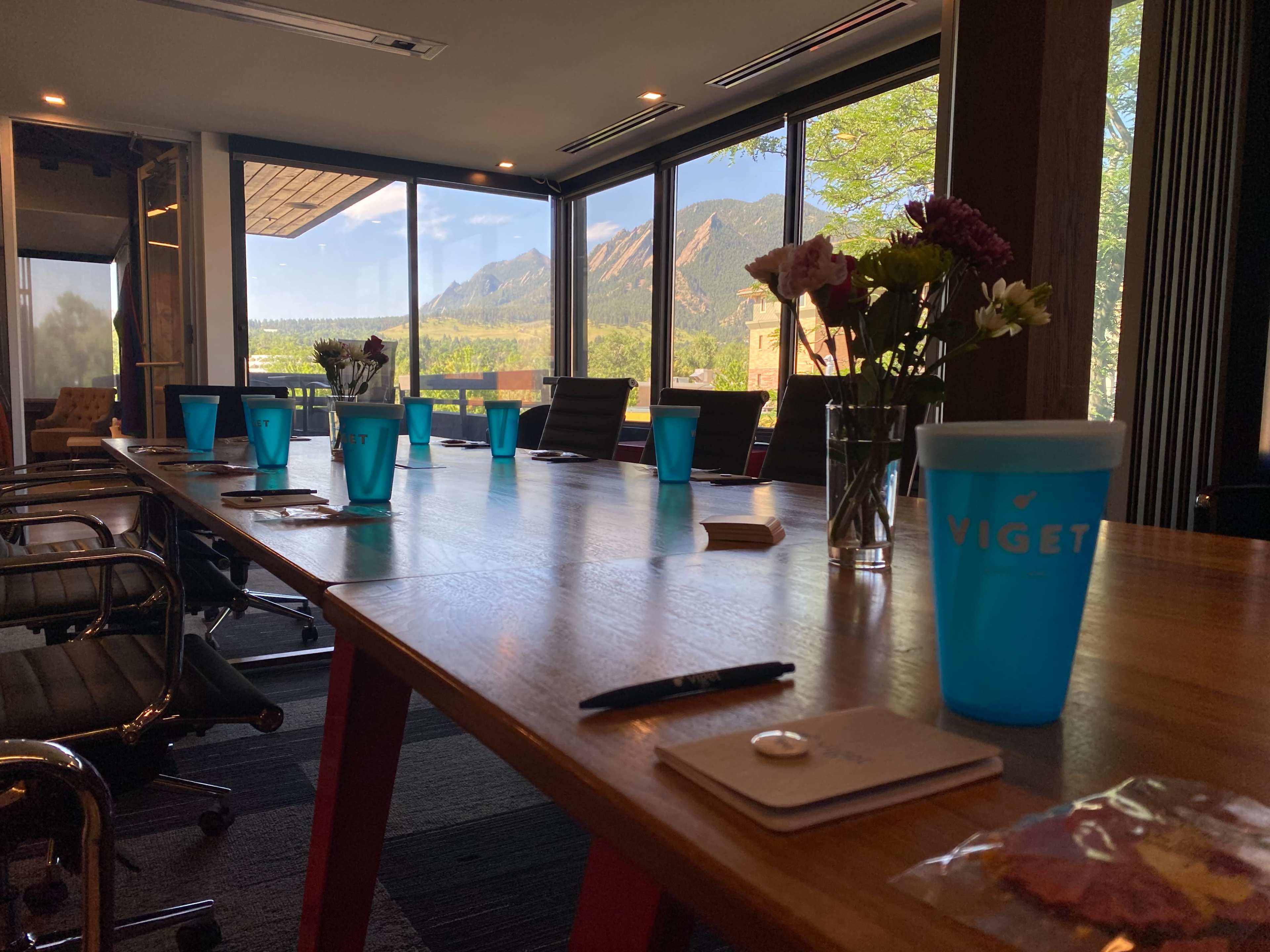 A conference room features a long wooden table set with blue cups, notepads, and a pen, overlooking a scenic view of mountains through large windows.