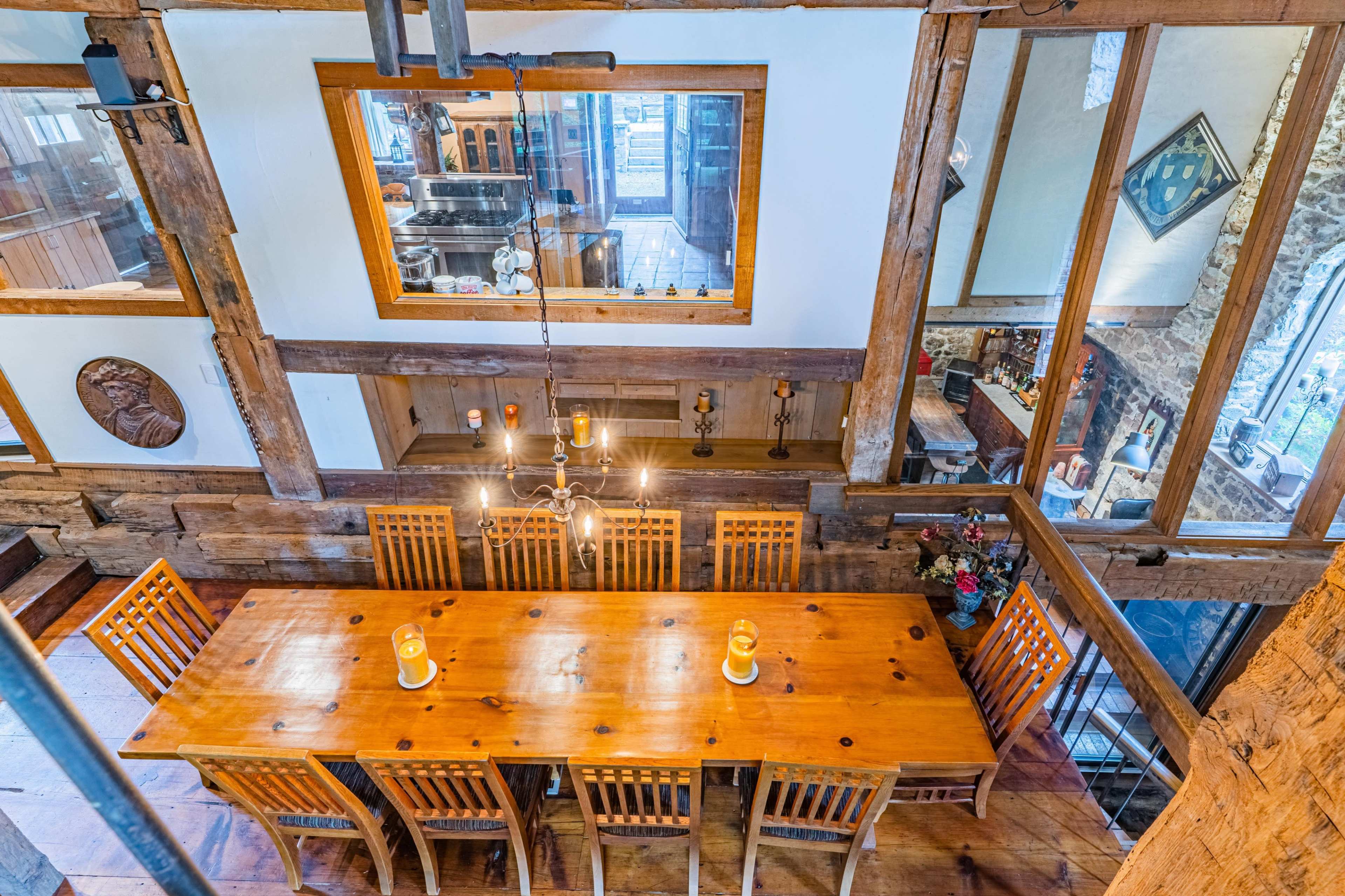 The image shows a spacious dining area with a long wooden table surrounded by chairs, set in a rustic room featuring wooden beams and large windows.