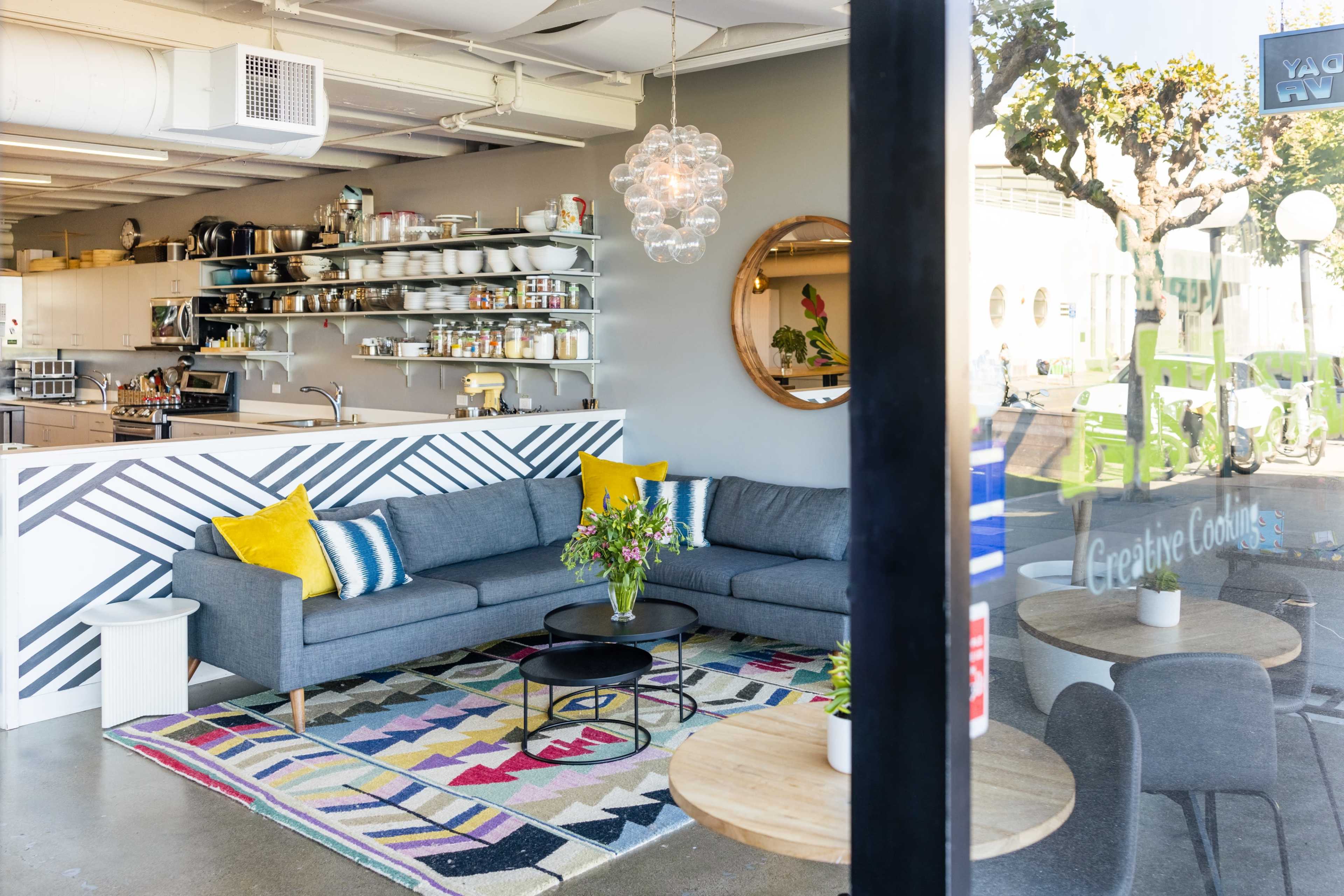 The scene shows a cozy, modern lounge area with a gray sectional sofa, a round coffee table, colorful patterned rugs, and shelves filled with kitchenware in a bright setting.
